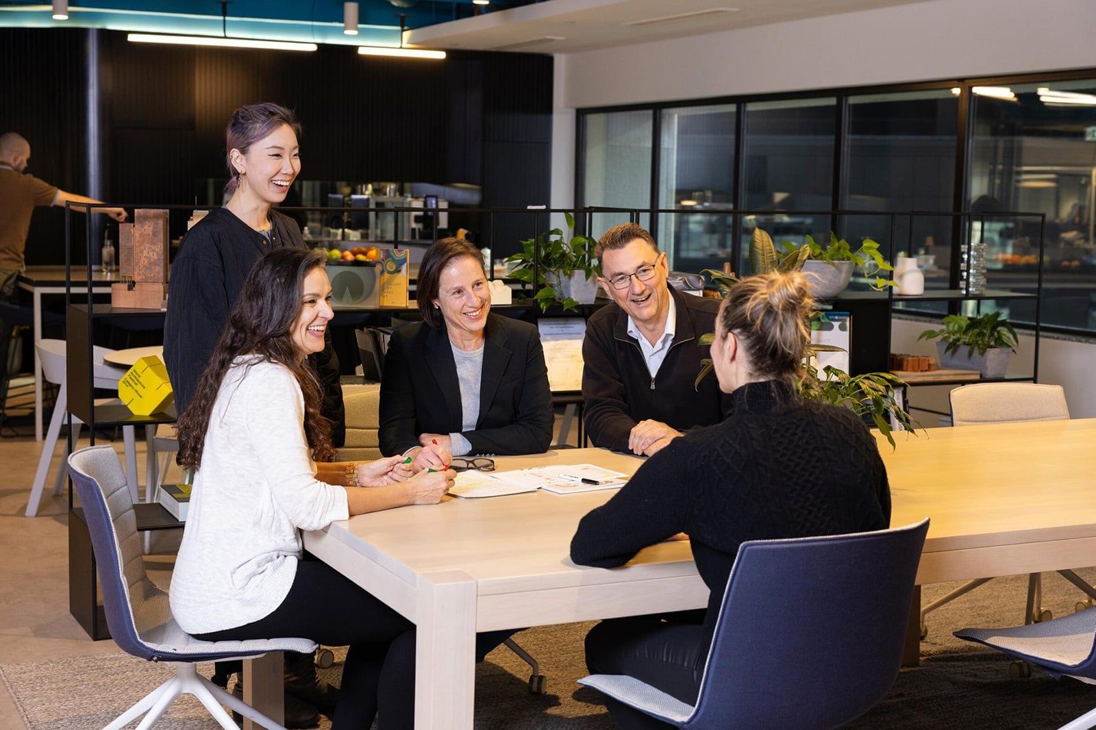 A group of five people smiling and having a meeting around a table in a modern office setting with plants in the background.