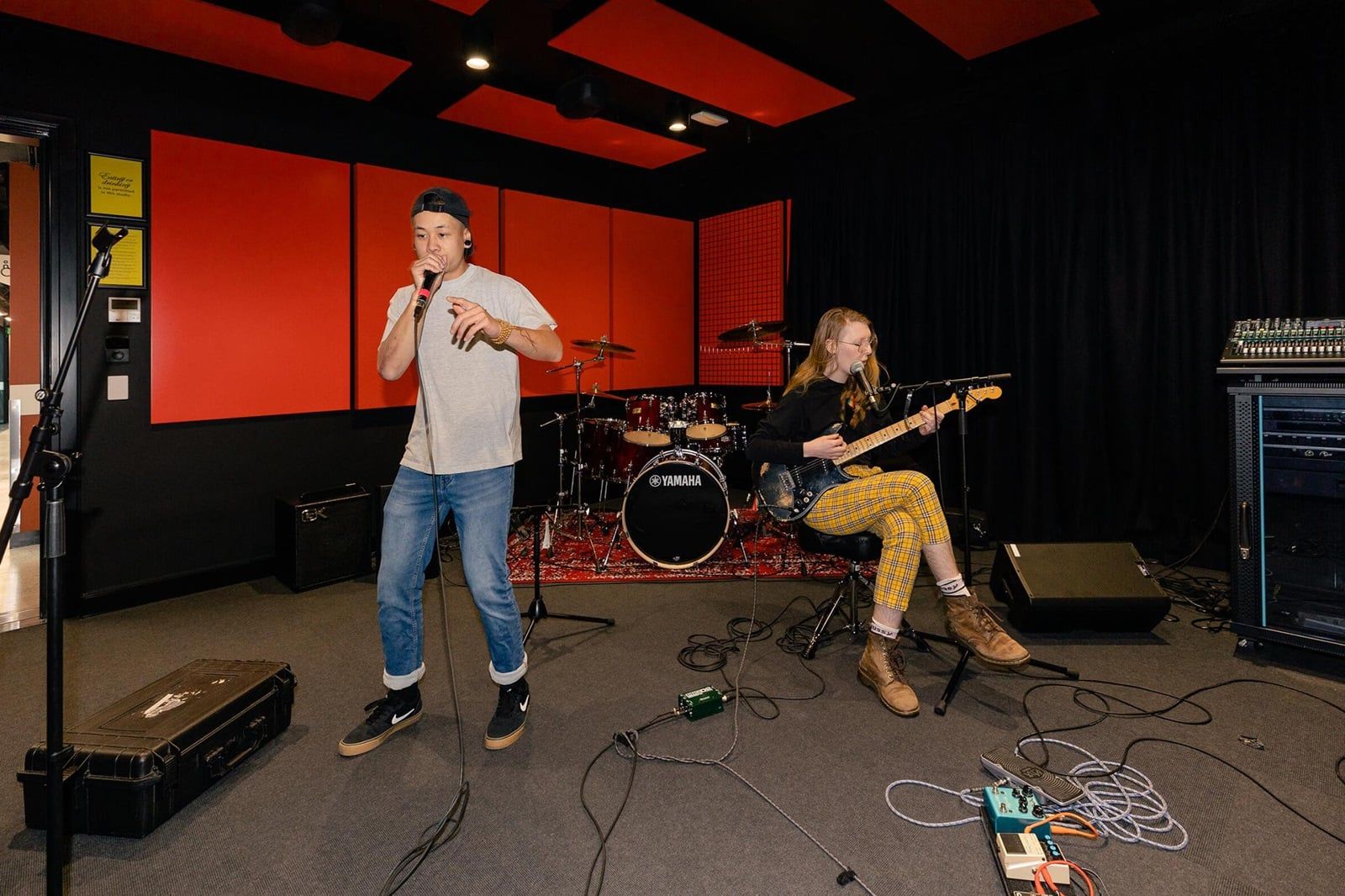 Two musicians in a studio: one singing with a microphone, the other playing an electric guitar, surrounded by musical equipment.