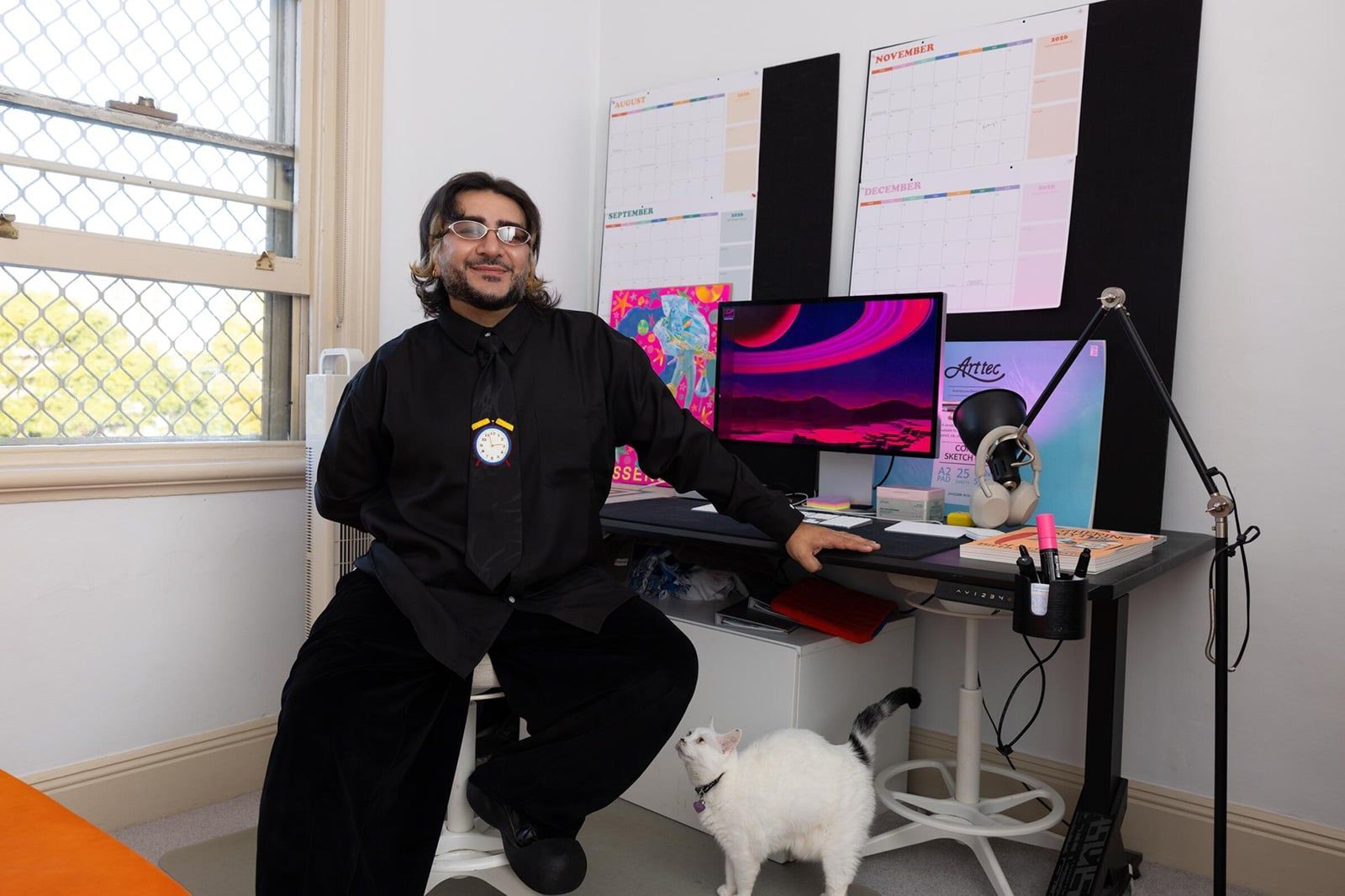 Person sitting on a desk, wearing a black shirt and sunglasses, with a white cat nearby. A colorful screen and planners are in the background.