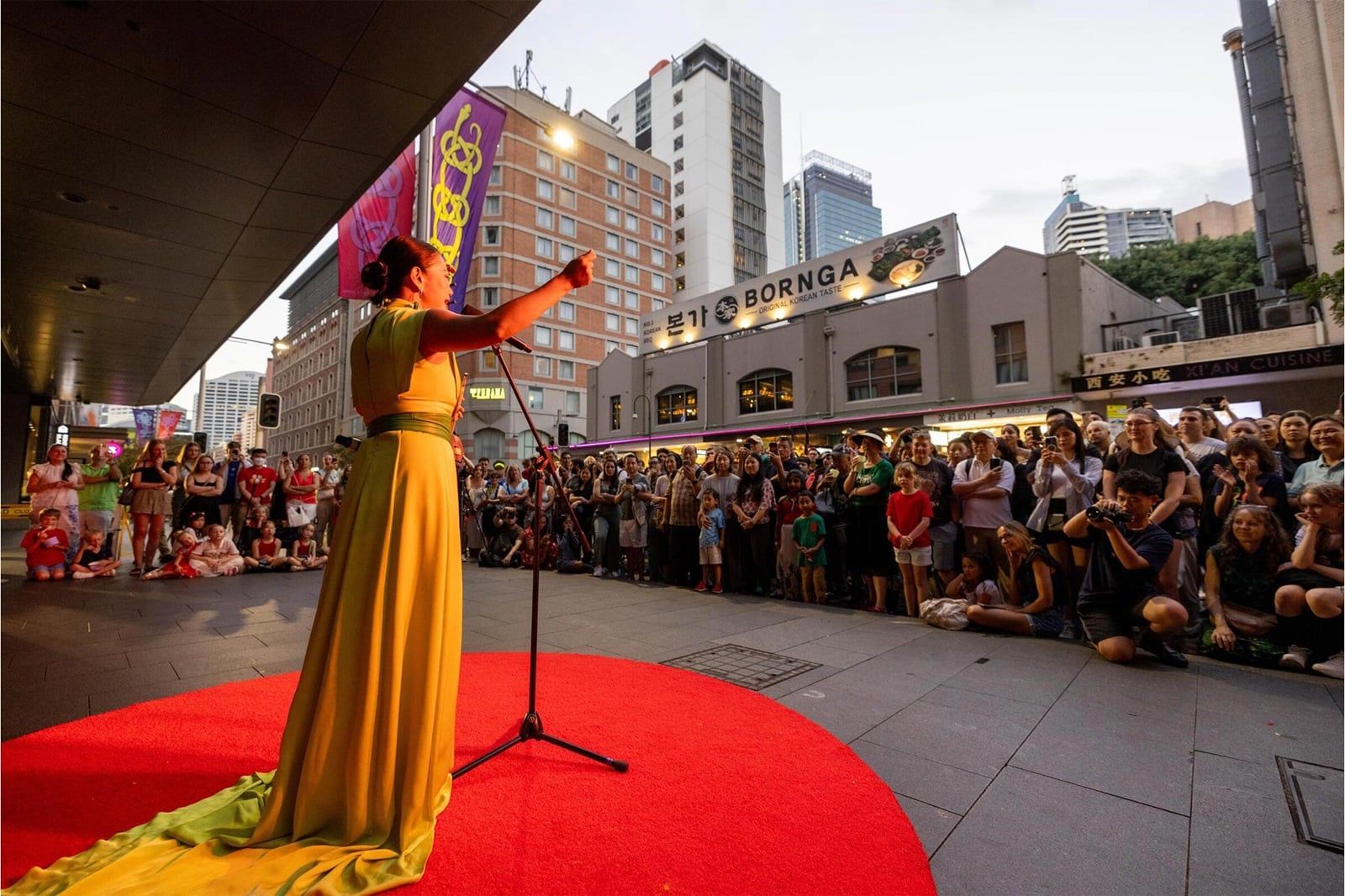 Performer in a yellow dress sings on a red carpet outside, with a large audience gathered under city buildings at dusk.