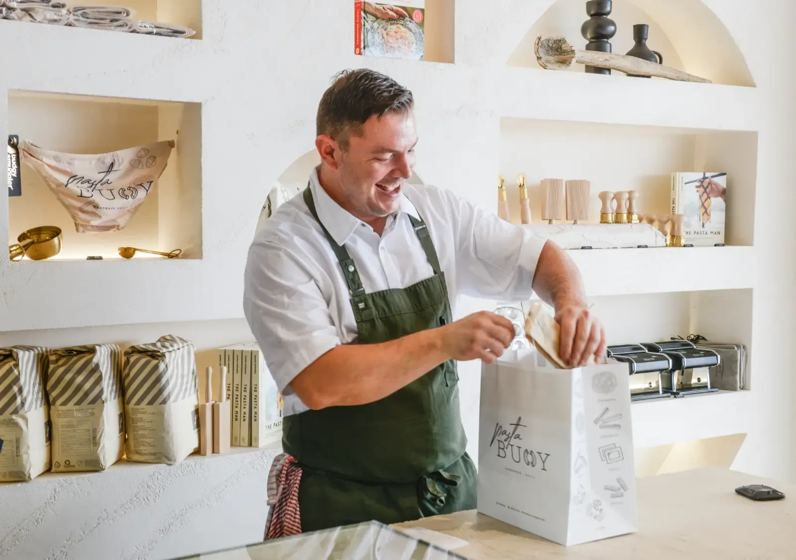 Smiling man in a green apron bags items in a chic store filled with pasta-themed products and kitchen tools.