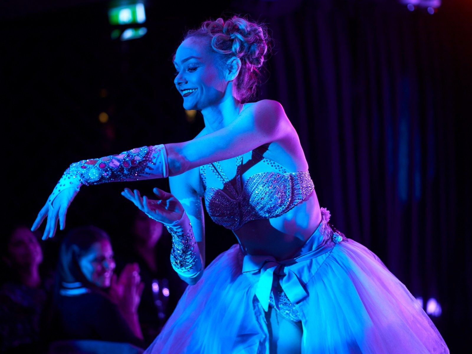 A smiling dancer wearing a tutu and bathed in blue light extends a graceful arm with a satin glove