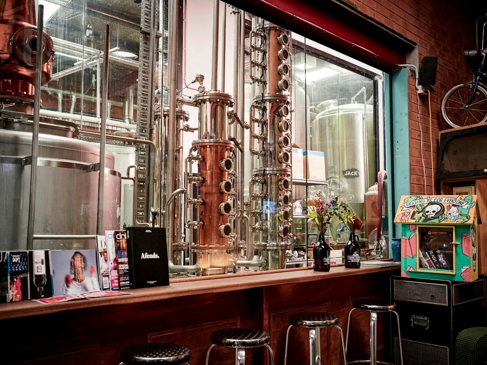 Distillery interior with copper machinery, stools by a wooden counter, magazines, flowers, and a vintage arcade game in the foreground.