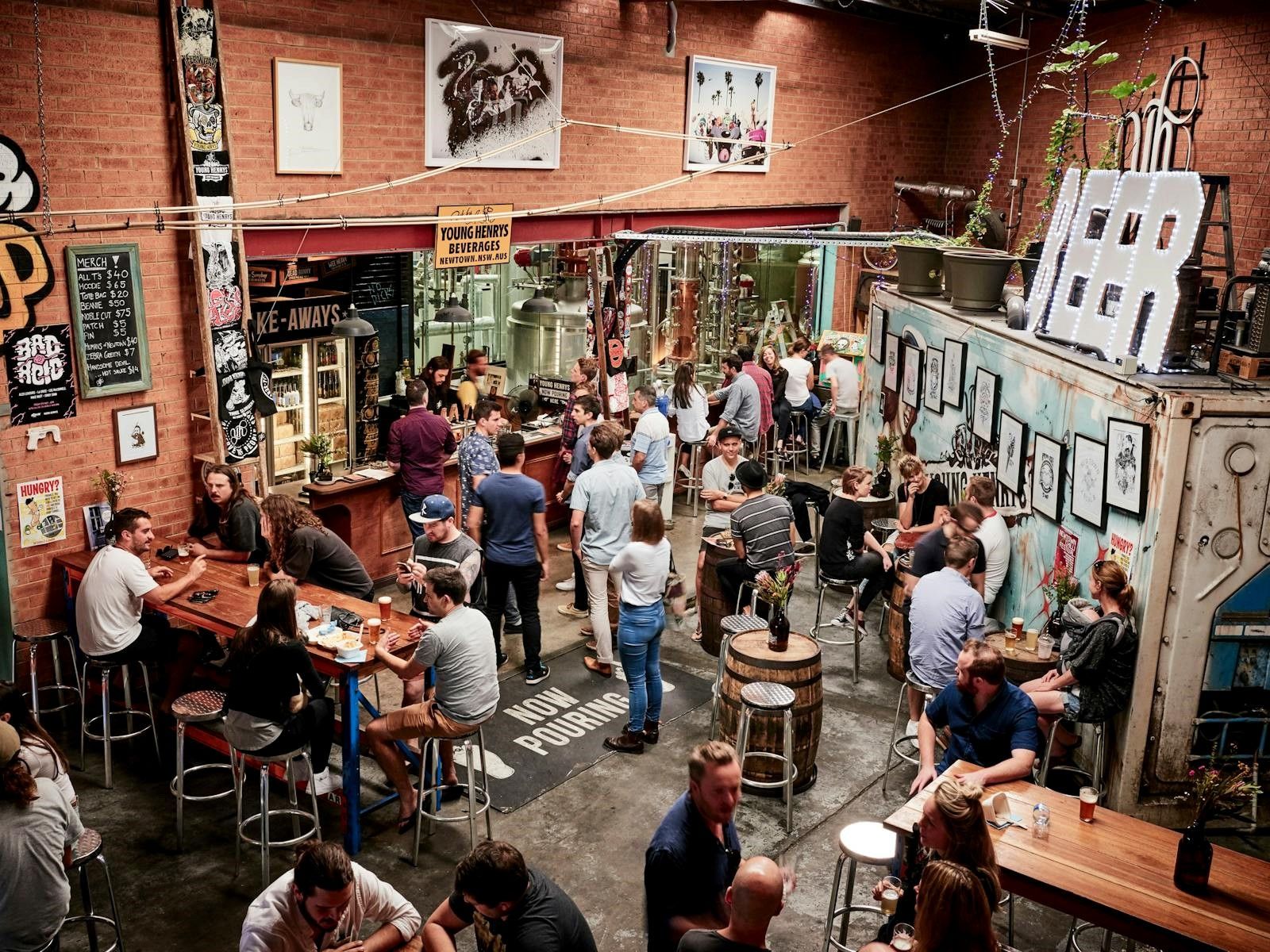 A lively brewery scene with people enjoying drinks at tables and the bar, surrounded by beer tanks, artwork, and a large "BEER" sign.