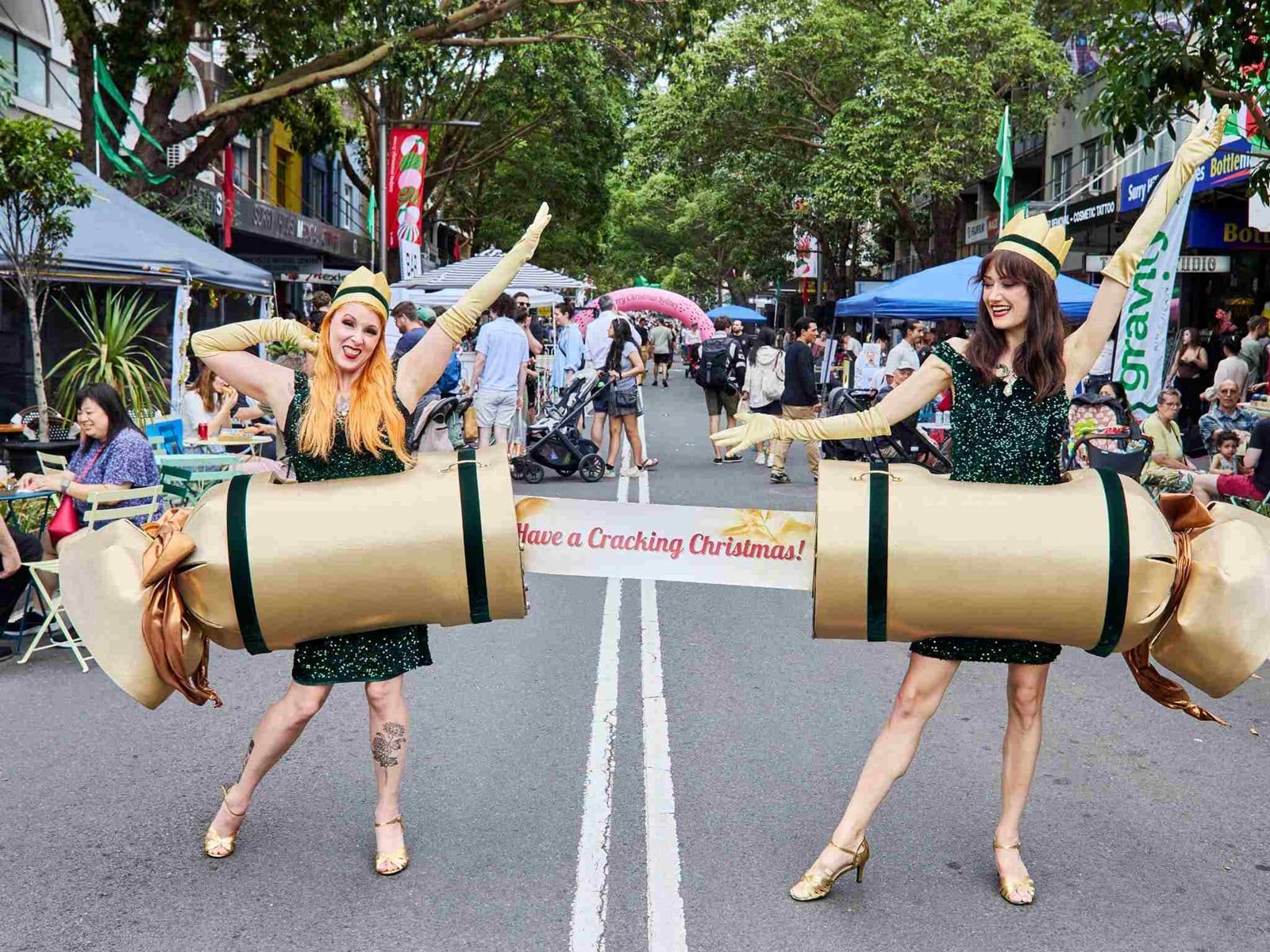 Two people in Christmas cracker costumes pose on a street with a "Have a Cracking Christmas!" banner amidst a festive outdoor market.