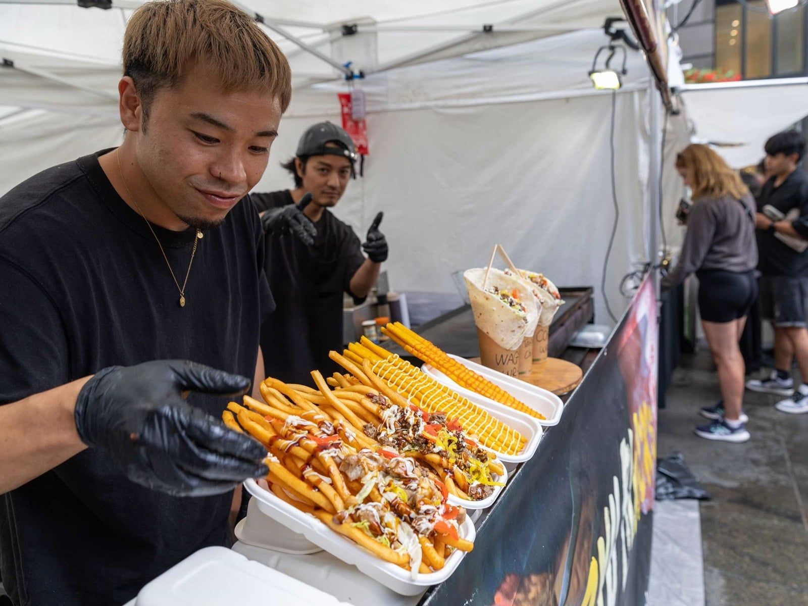 Vendor serving loaded fries and skewered corn at a food stall, with people lined up in the background.