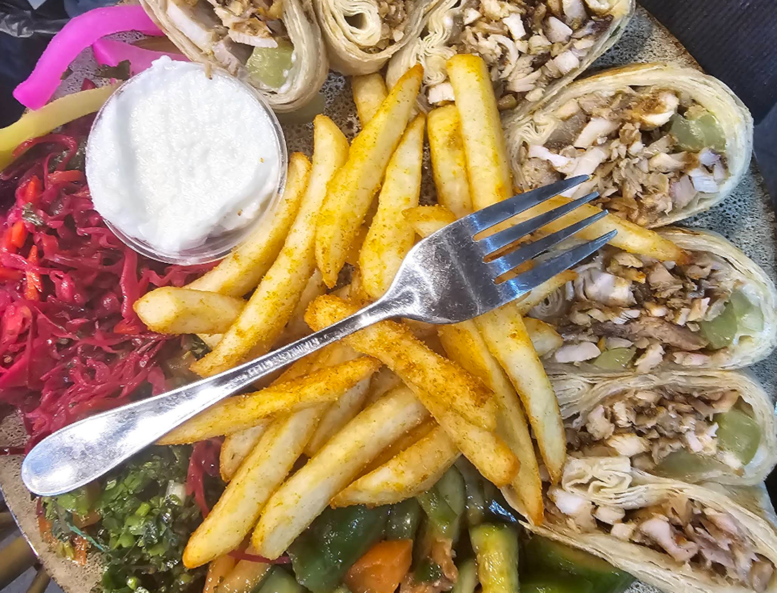A plate with sliced wraps, seasoned fries, pickled vegetables, shredded cabbage, and a small cup of white sauce, with a fork on top.