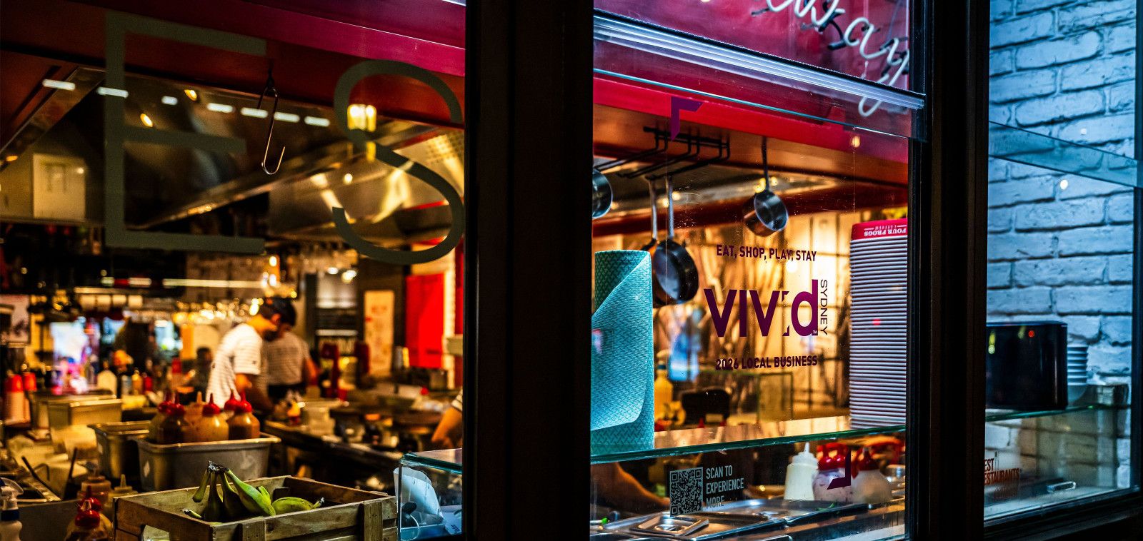View through a restaurant window showing a bustling kitchen with hanging utensils and a sign promoting "Vivid Sydney."