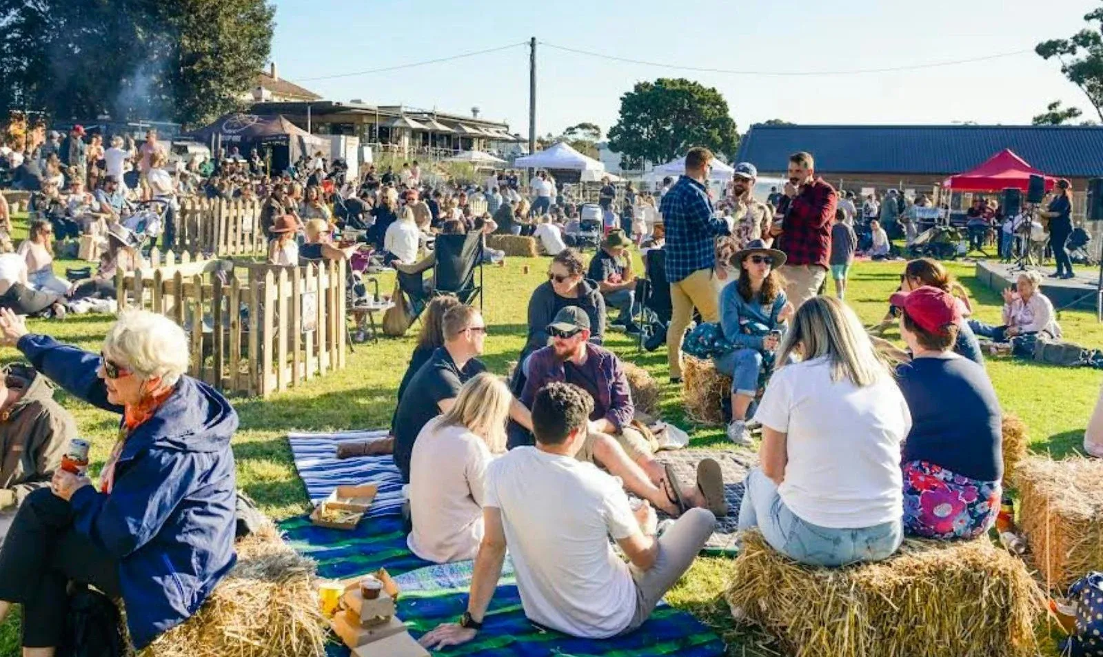 People enjoying an outdoor festival, sitting on hay bales and blankets in a sunny field, with food, drinks, and live music in the background.