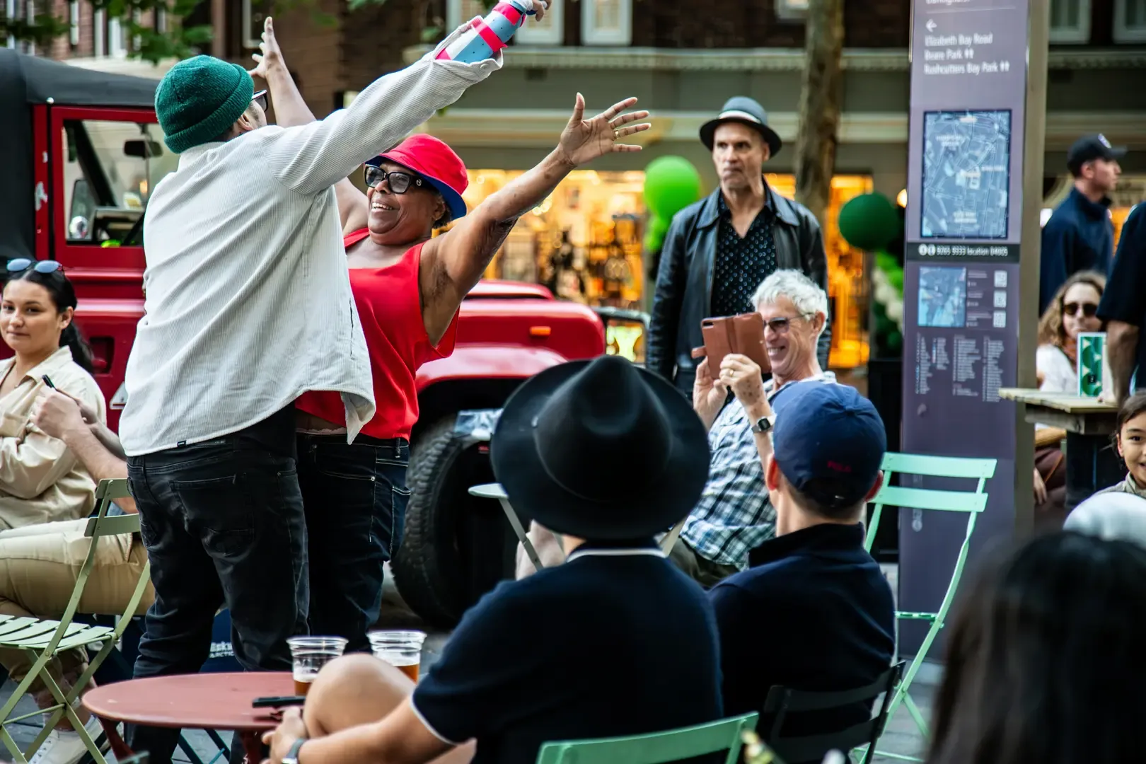 Two people joyfully high-fiving outdoors, surrounded by seated onlookers. A red vehicle and a map sign are visible in the background.