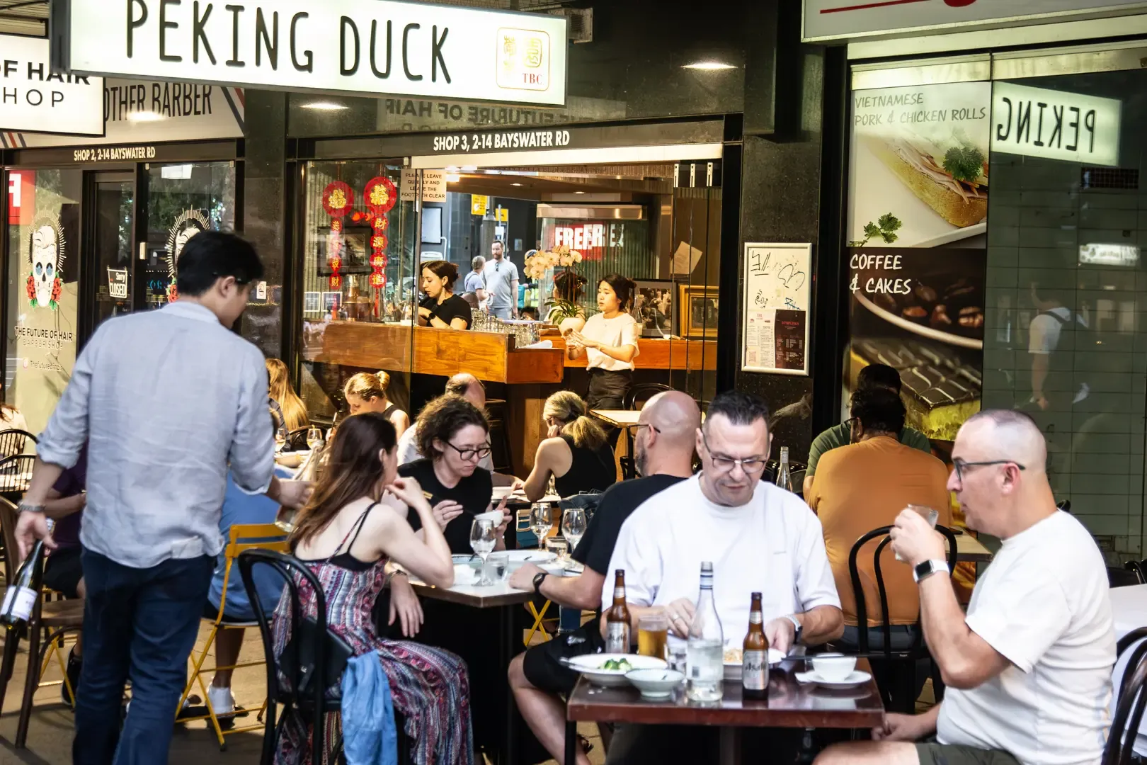People dining at outdoor tables of a bustling restaurant named "Peking Duck" at night, with a view of the busy interior through large windows.