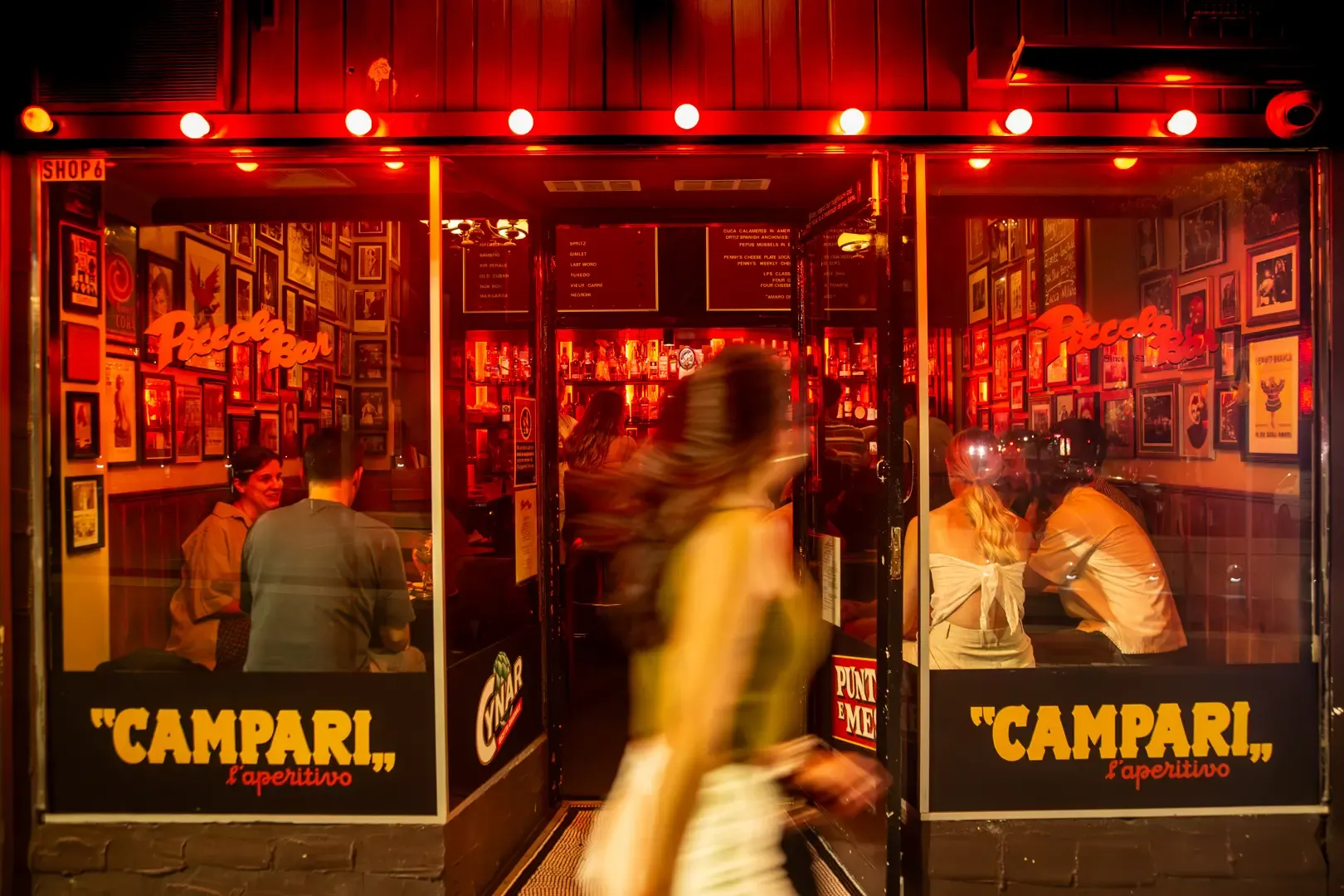 A woman walks past a lively, warmly-lit bar with people inside. The windows display "Campari" and are adorned with photos and posters.