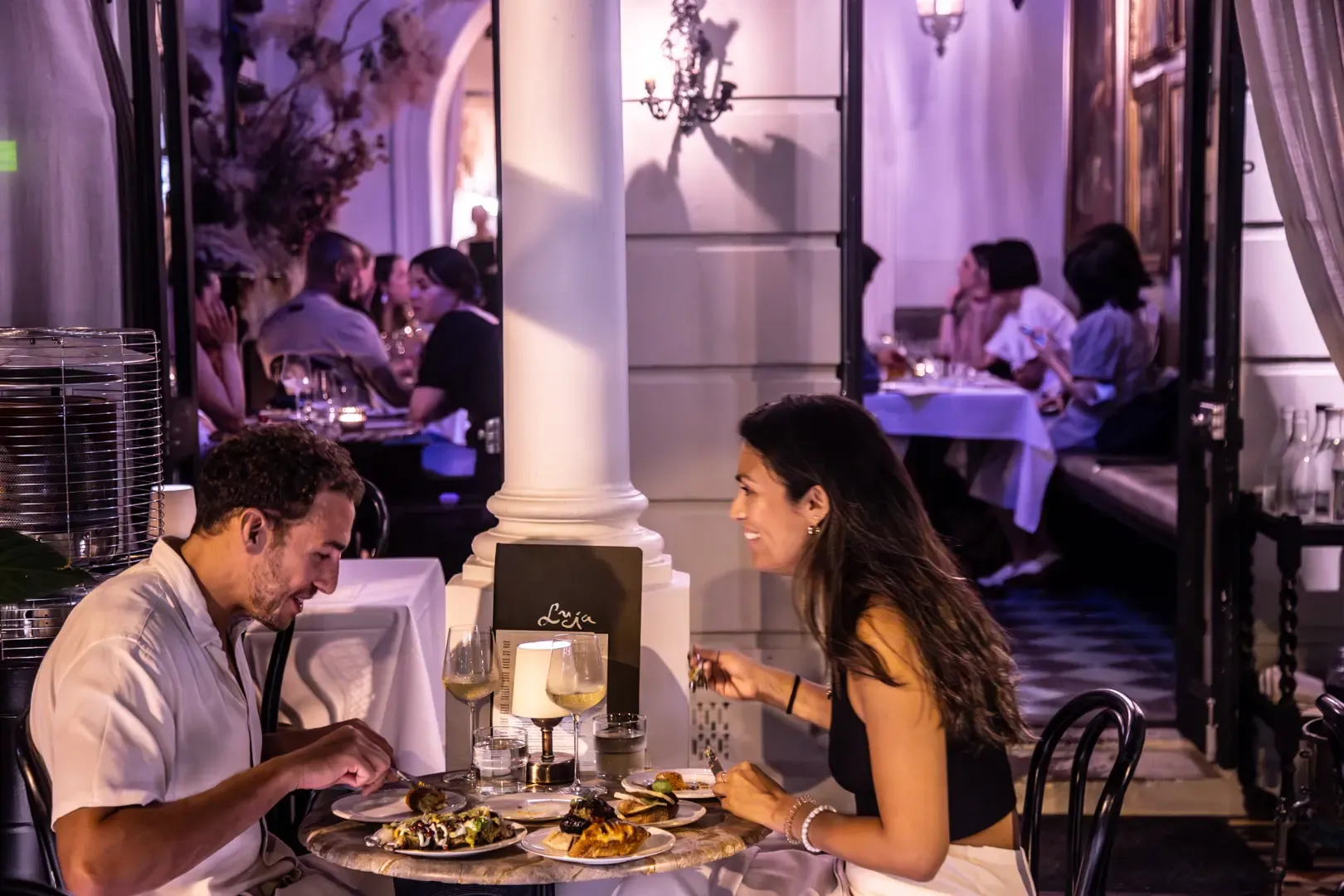 A couple enjoys dinner at an elegant, dimly lit restaurant. Other patrons are seated in the background, with soft lighting and ornate decor.