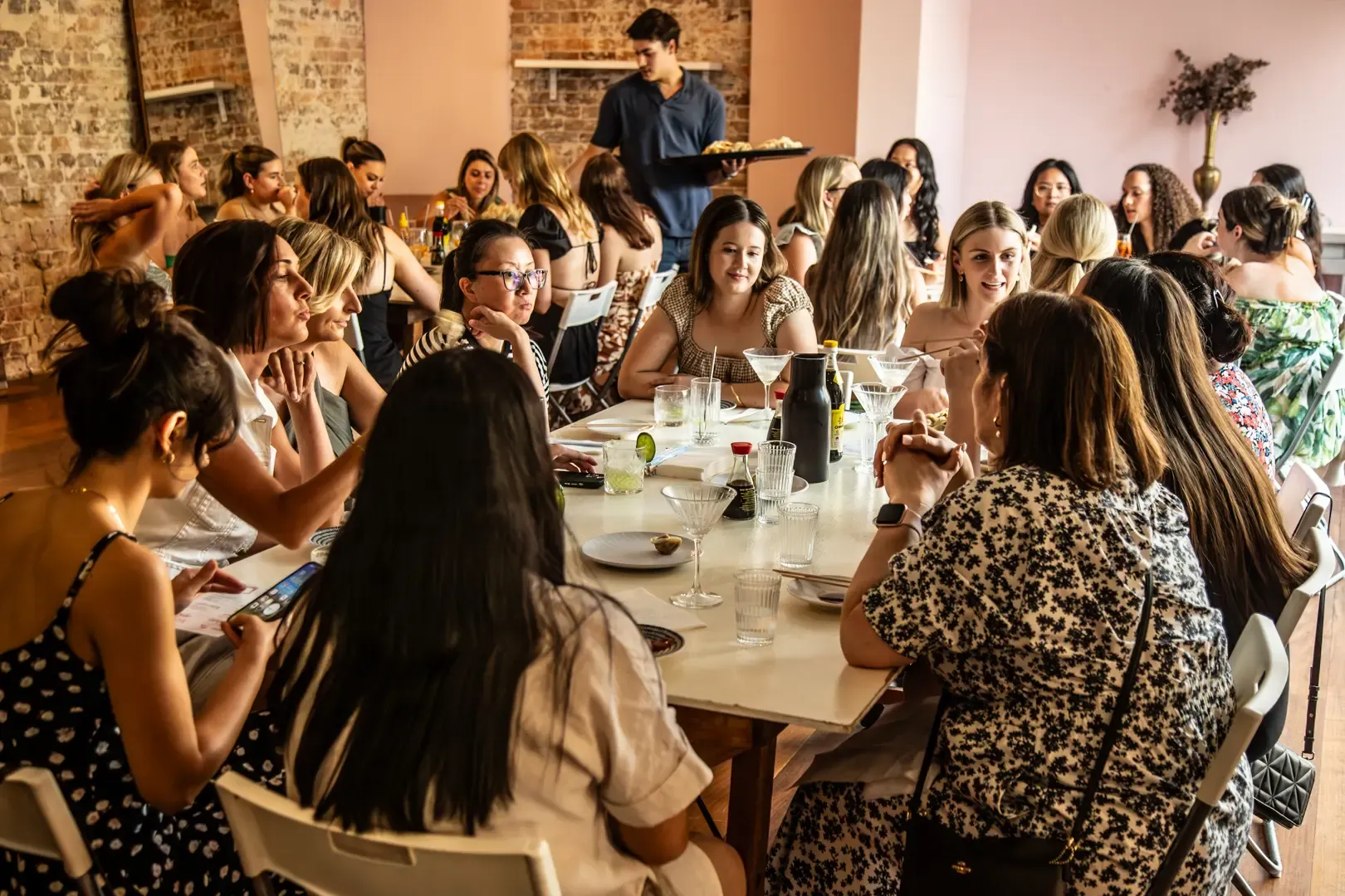 A group of people seated around a long table in a restaurant, engaged in conversation and dining. A waiter serves food in the background.