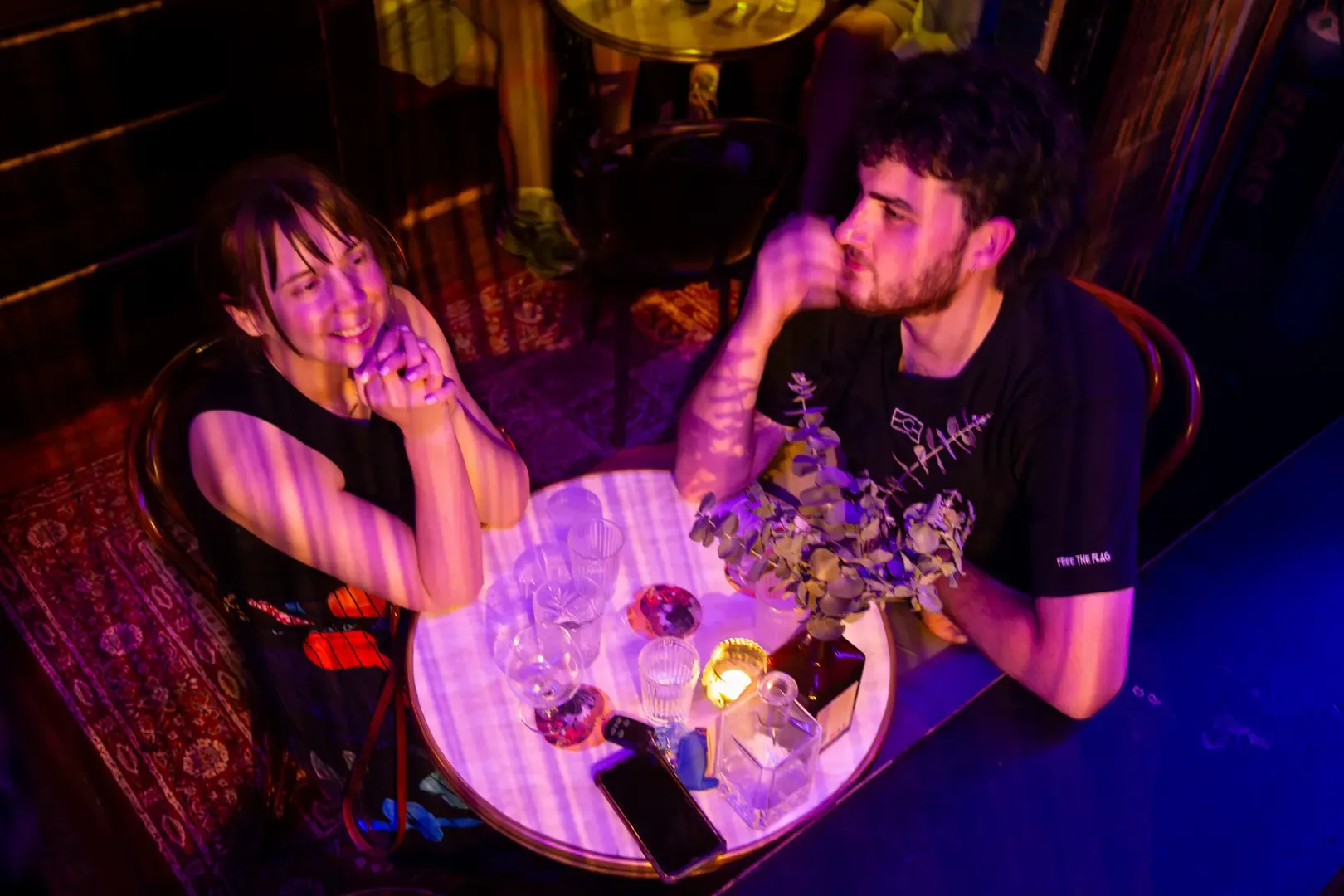 A woman and a man sit at a small round table with drinks and a candle, smiling and talking in a dimly lit, cozy setting.