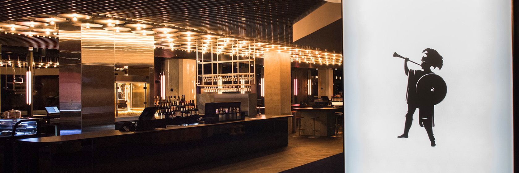 Dimly lit modern bar interior with reflective surfaces, featuring a silhouetted figure playing a trumpet on a nearby wall.