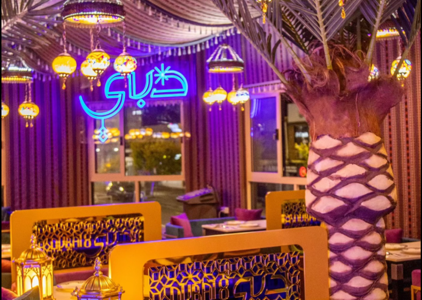 Colorful Middle Eastern restaurant interior with hanging lanterns, a decorative palm tree, and a neon sign in Arabic script.