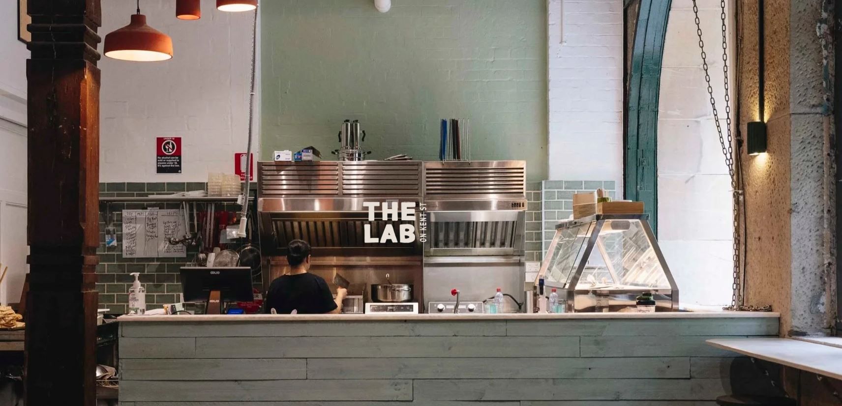 A person works in a modern, industrial-style kitchen labeled "The Lab," with stainless steel appliances and green tiled walls.