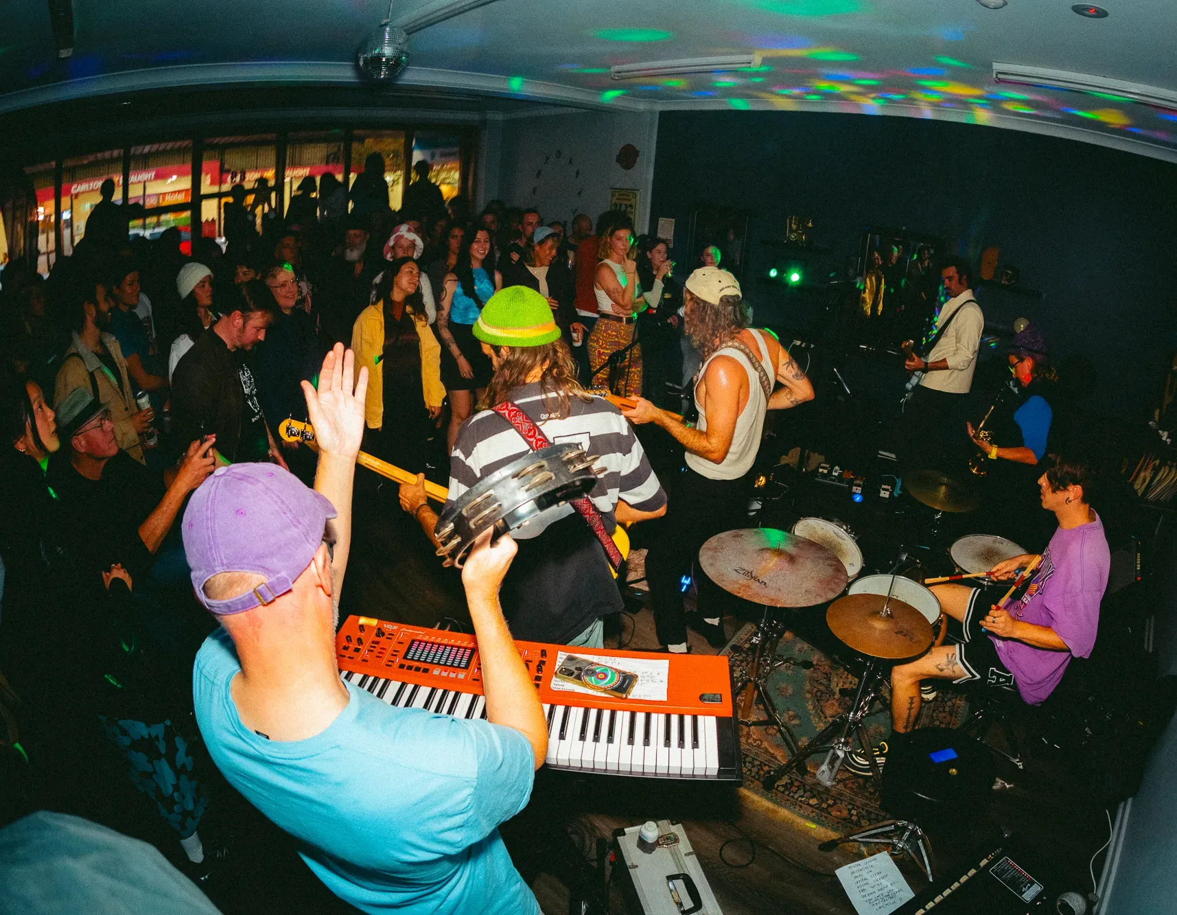 Band performing indoors with musicians on keyboards, drums, and guitar. Colorful lights and a lively audience create a vibrant atmosphere.
