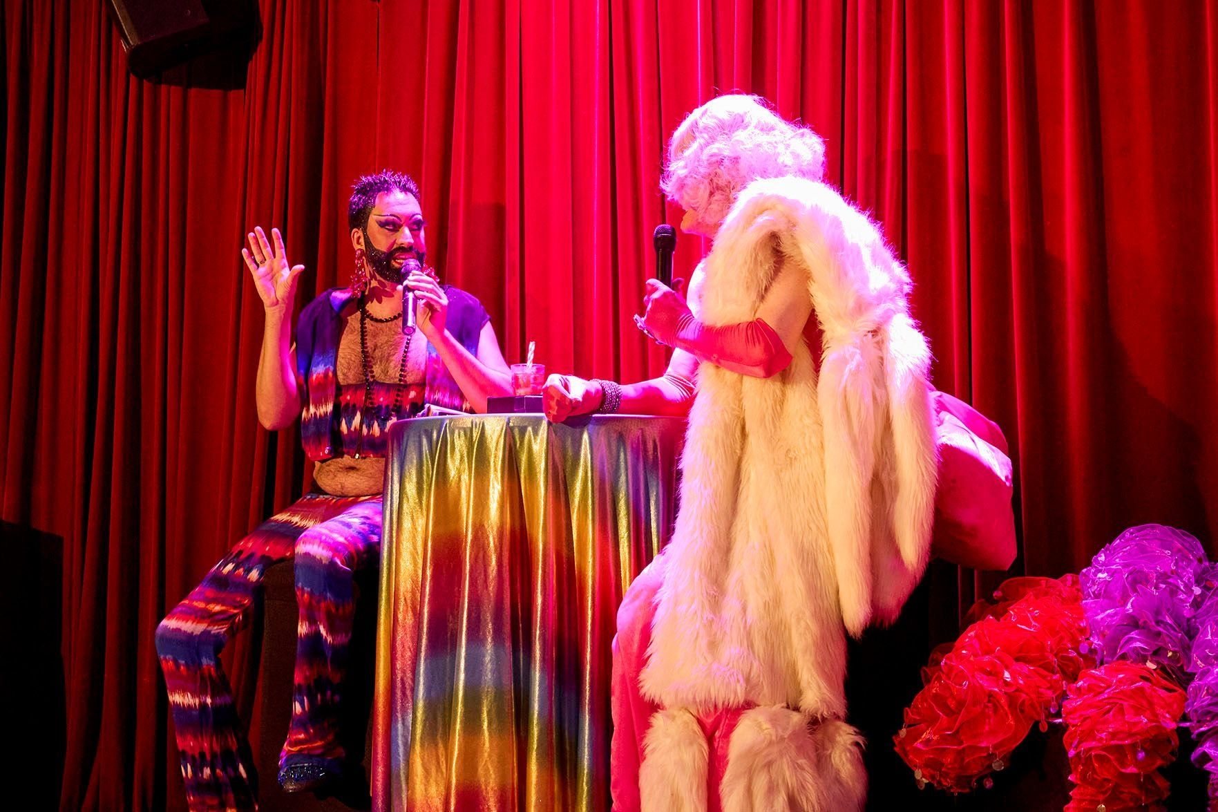 Two performers in vibrant costumes and makeup sit at a table with a rainbow cloth, engaging in conversation on a stage with red curtains.