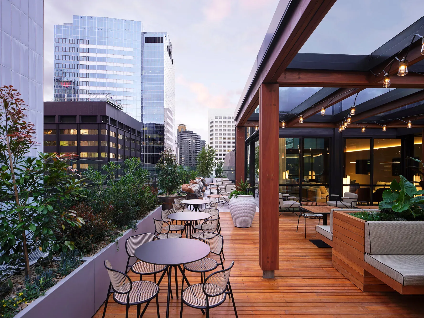 Rooftop seating area with modern furniture, wooden deck, and plants, surrounded by tall glass buildings under a cloudy sky.