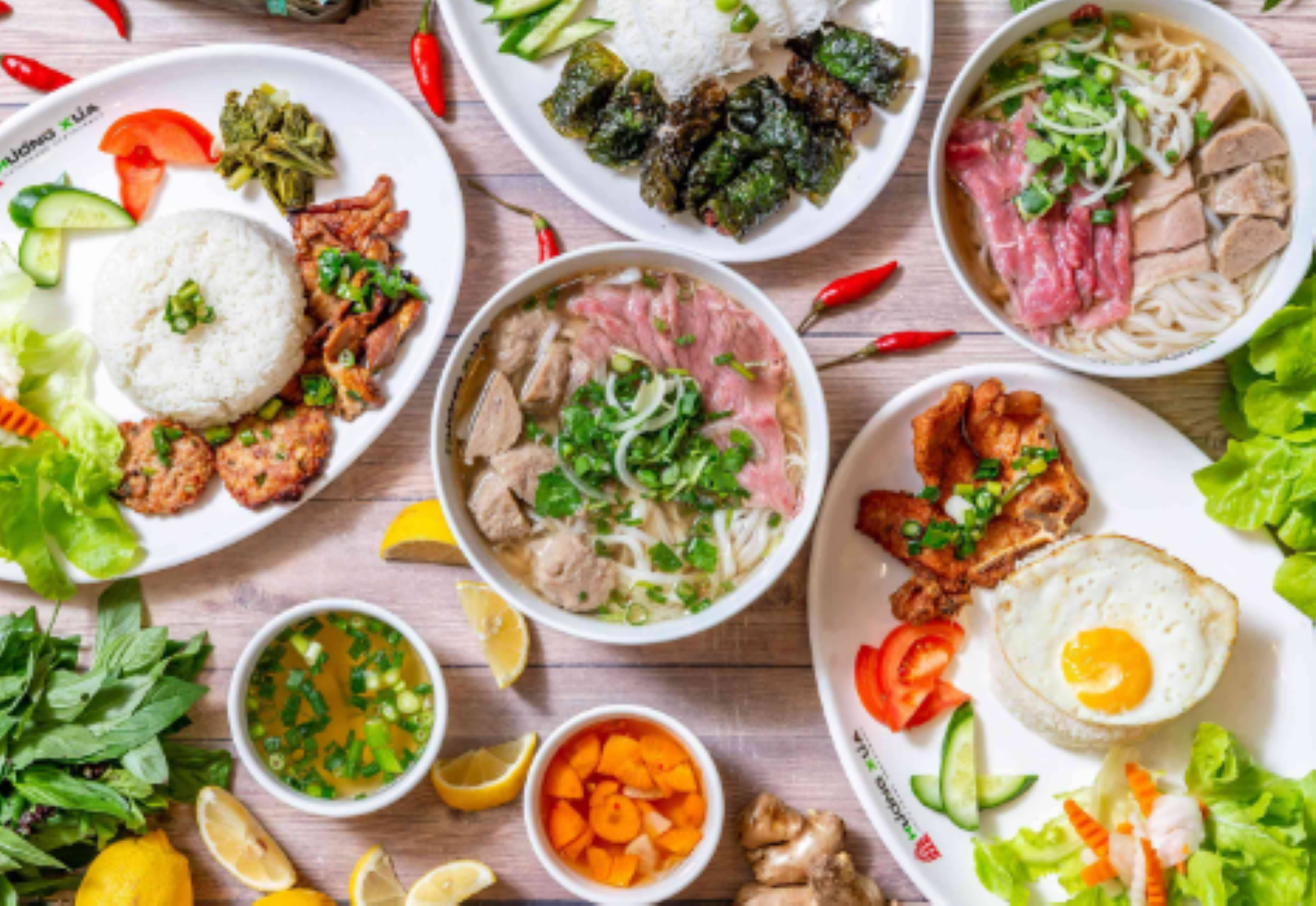 A variety of Vietnamese dishes, including pho, rice with sides, fresh herbs, and garnishes, arranged on a wooden table.