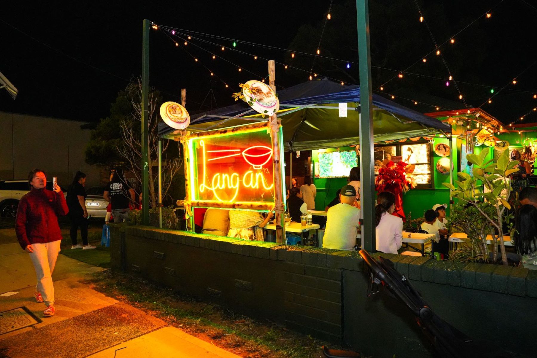 Outdoor eatery at night with a glowing "Lang an" neon sign, string lights, and diners seated at tables under a tent.