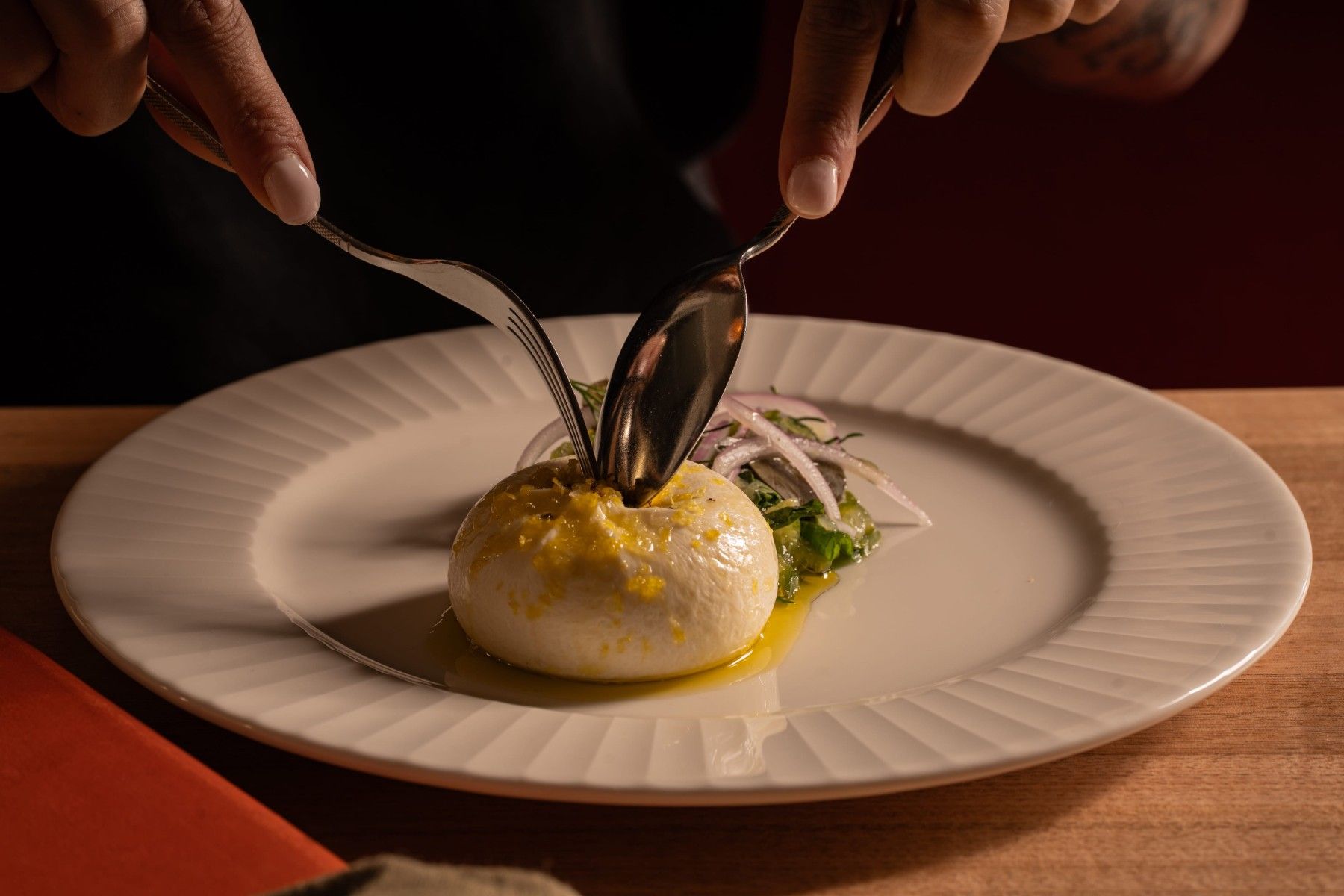 Person using spoons to open a burrata cheese on a plate with sliced onions and greens on the side, placed on a wooden table.