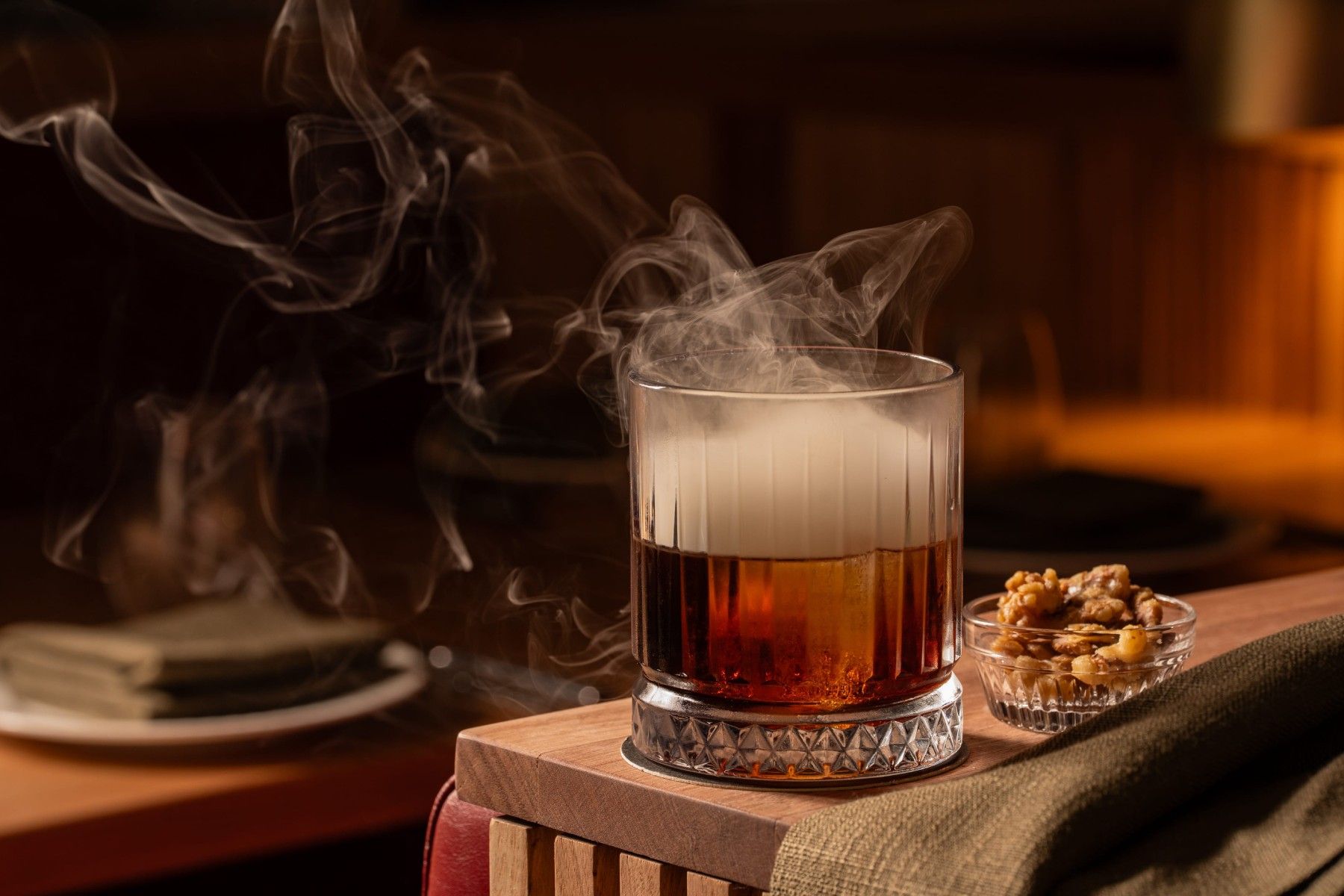 A smoky cocktail in a glass with a textured base, next to a small bowl of mixed nuts on a wooden table.