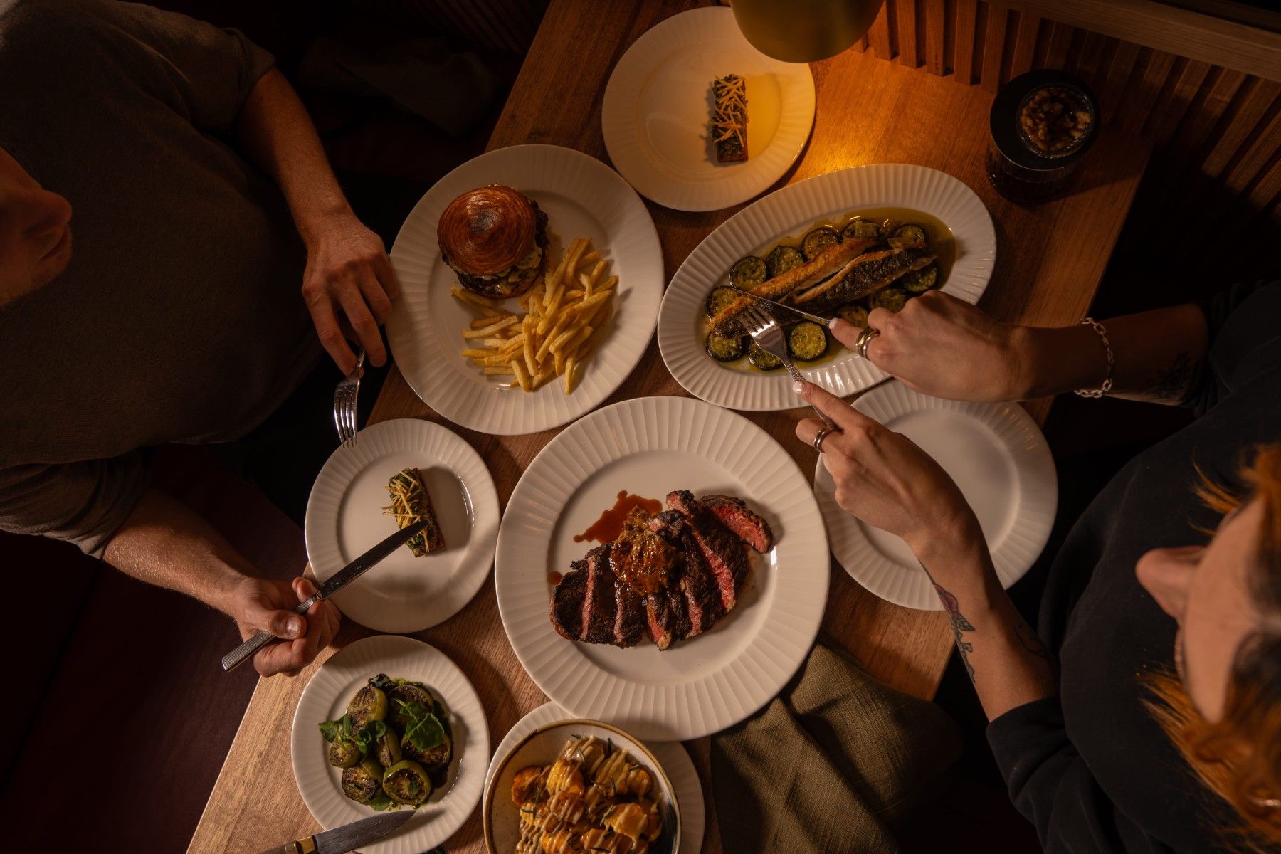 Three people dining at a table with plates of steak, fries, burger, salad, and a taco, viewed from above in warm lighting.