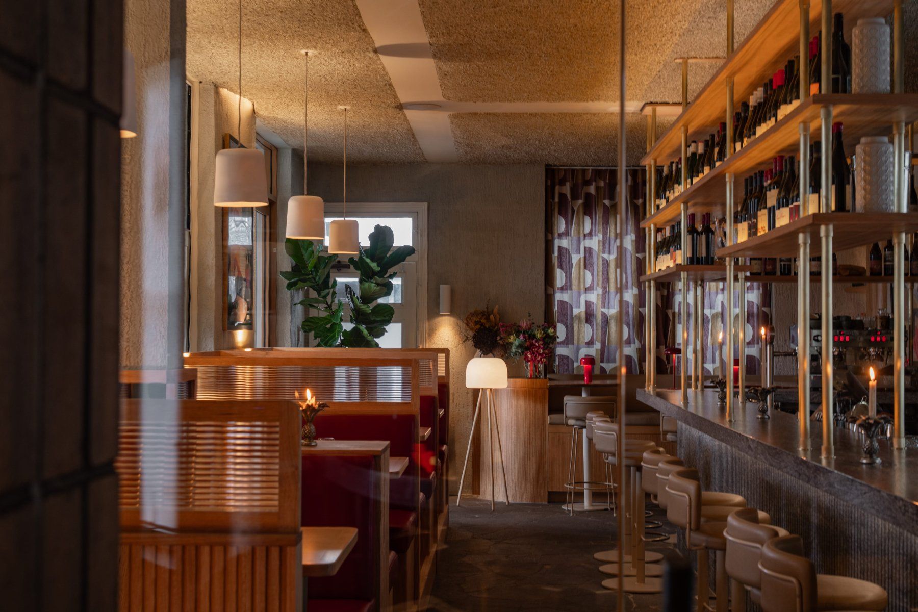 Cosy restaurant interior with wooden booths, a bar with stools, dim lighting, and decorative plants. Shelves display wine bottles.