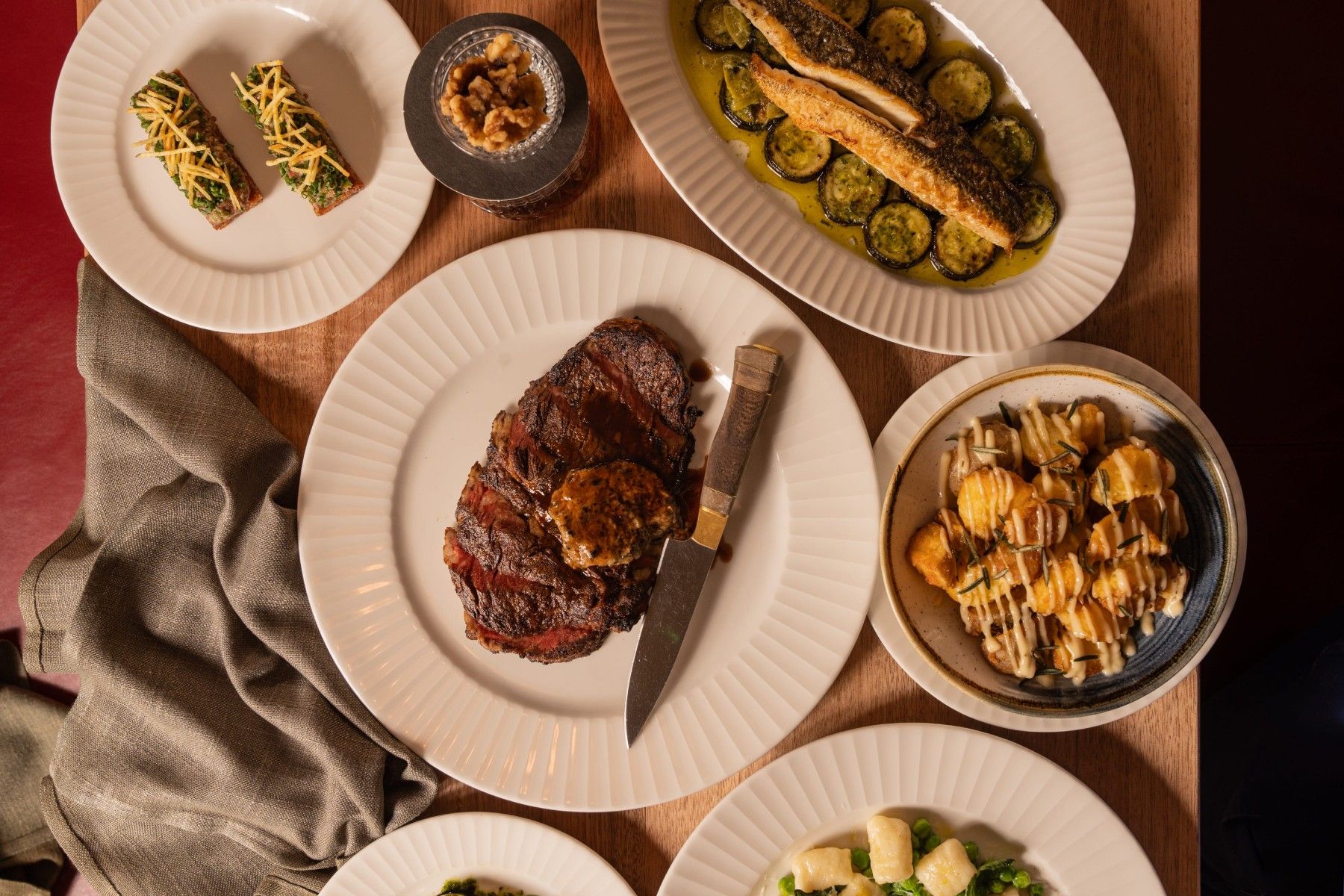 An overhead view of a table with a steak, zucchini, fish, gnocchi, and appetizers on white plates, with a knife and napkin.