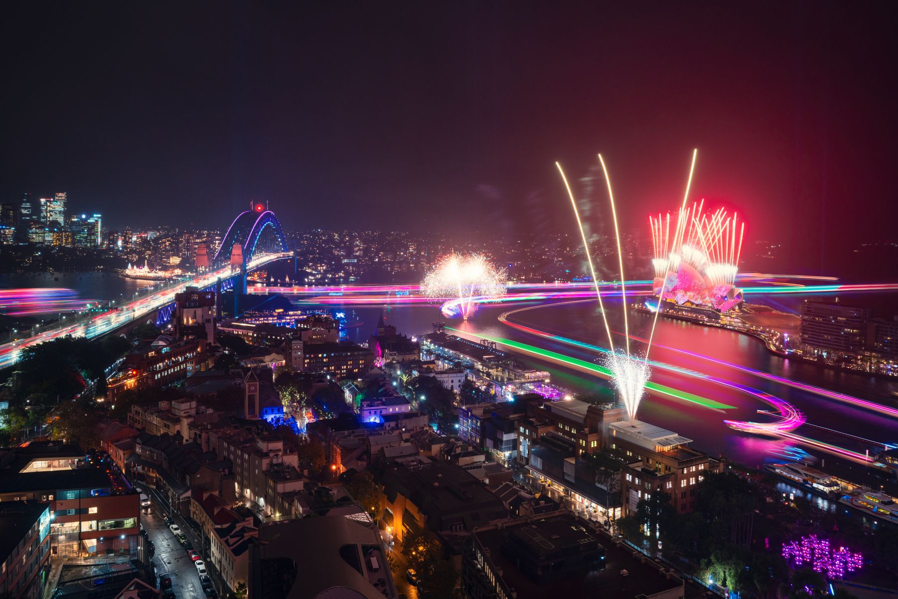 A vibrant night view of a city harbour with colourful fireworks and light trails illuminating the sky and water, with a lit bridge in the background.