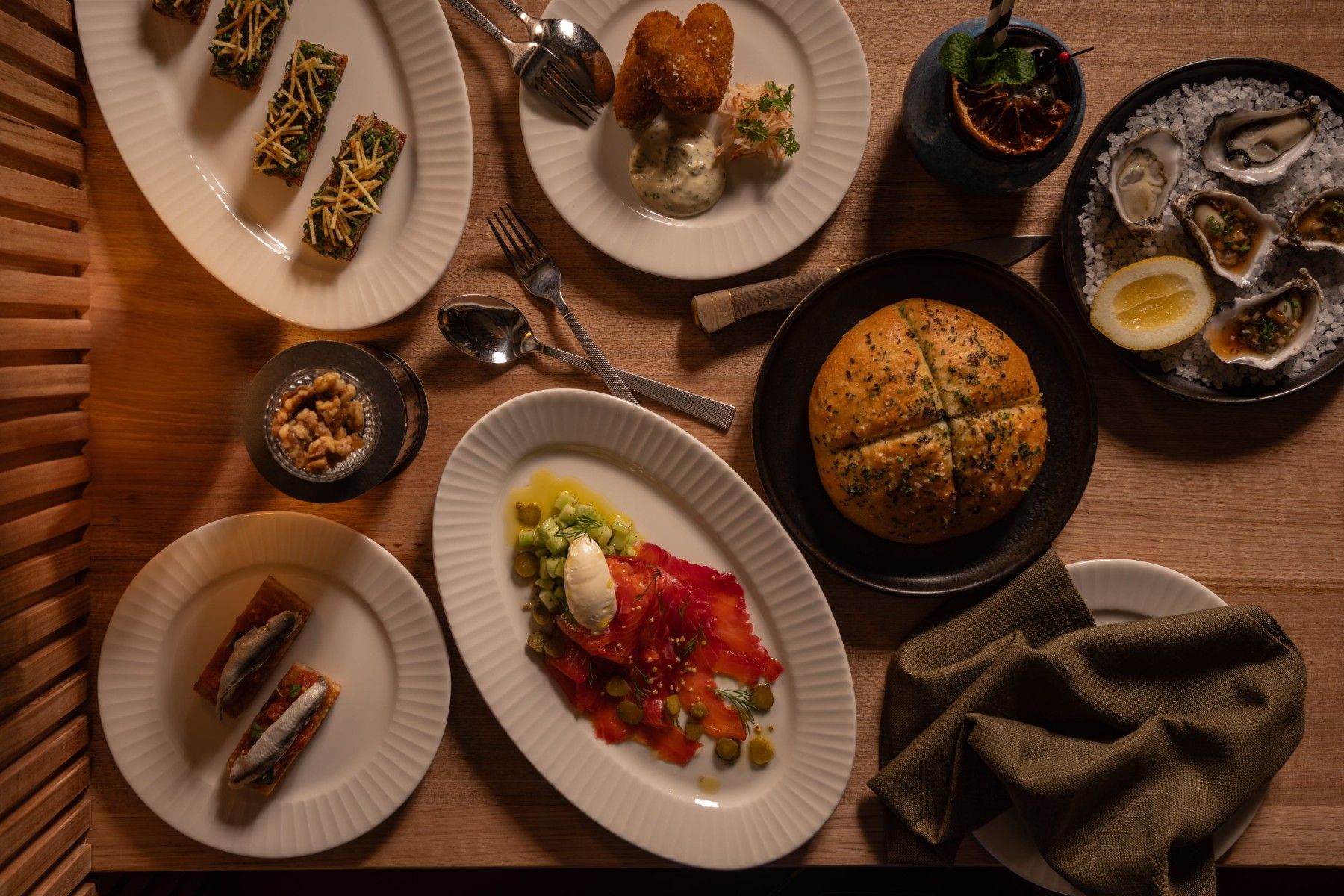 A wooden table with various dishes, including bread, oysters, fish, and desserts, elegantly arranged with cutlery and a green napkin.