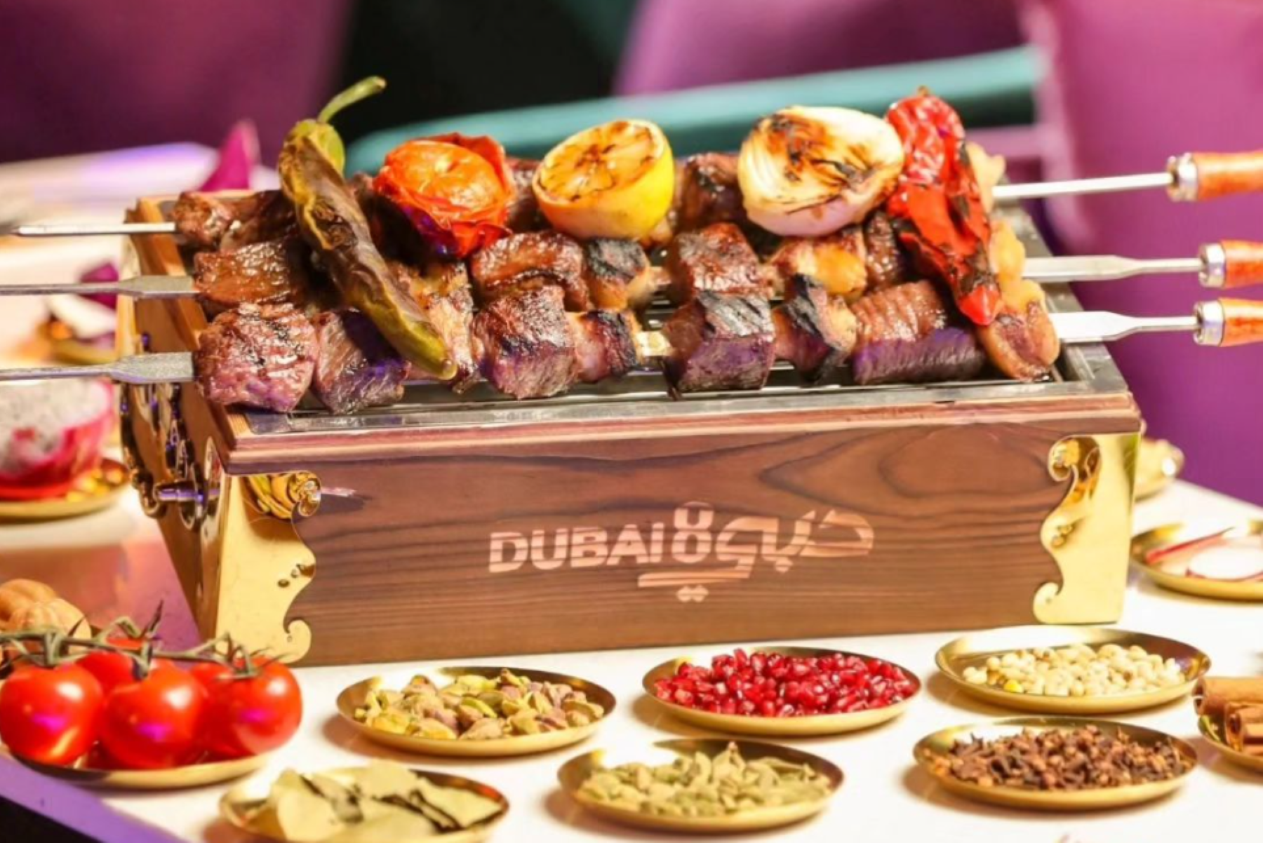 Skewers of grilled meat and vegetables on a wooden box with "Dubai8" written on it, surrounded by small bowls of spices and ingredients.