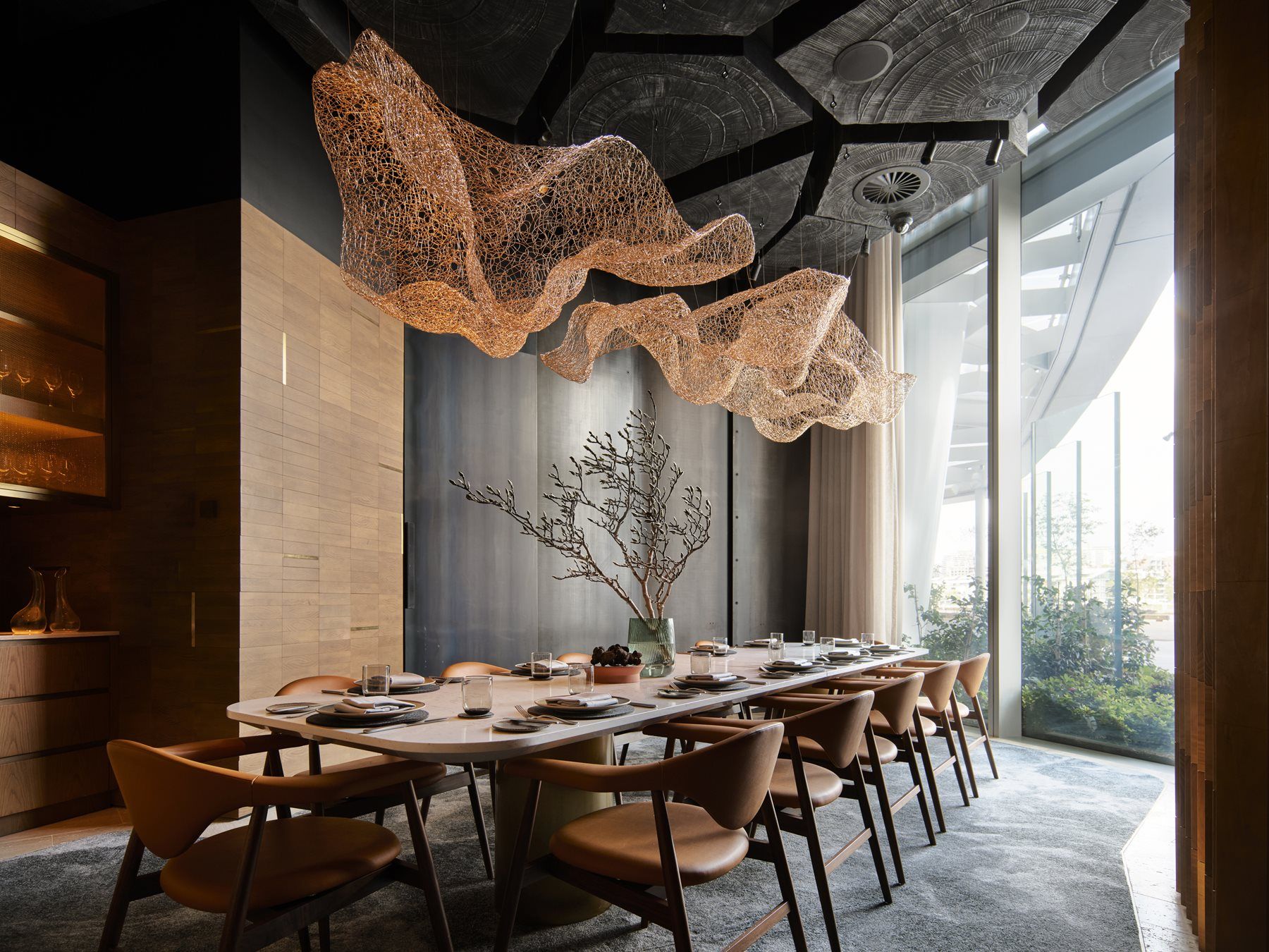 Elegant dining room with a long table, modern chairs, intricate ceiling art, minimalistic decor, and large windows overlooking greenery.