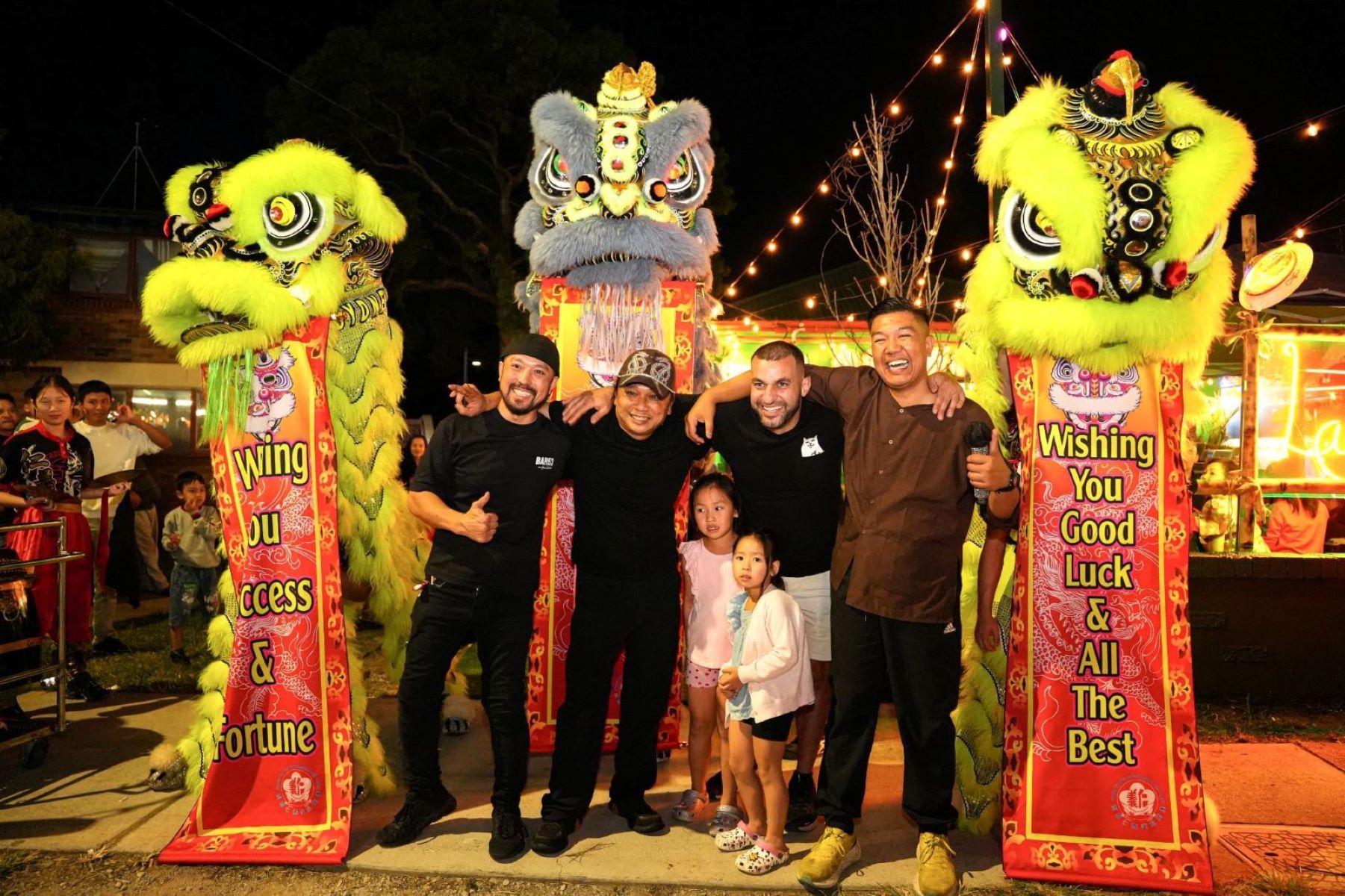 Group posing with colourful lion dance costumes and festival lights, banners reading "Wishing You Success & Fortune" and "Wishing You Good Luck & All The Best."