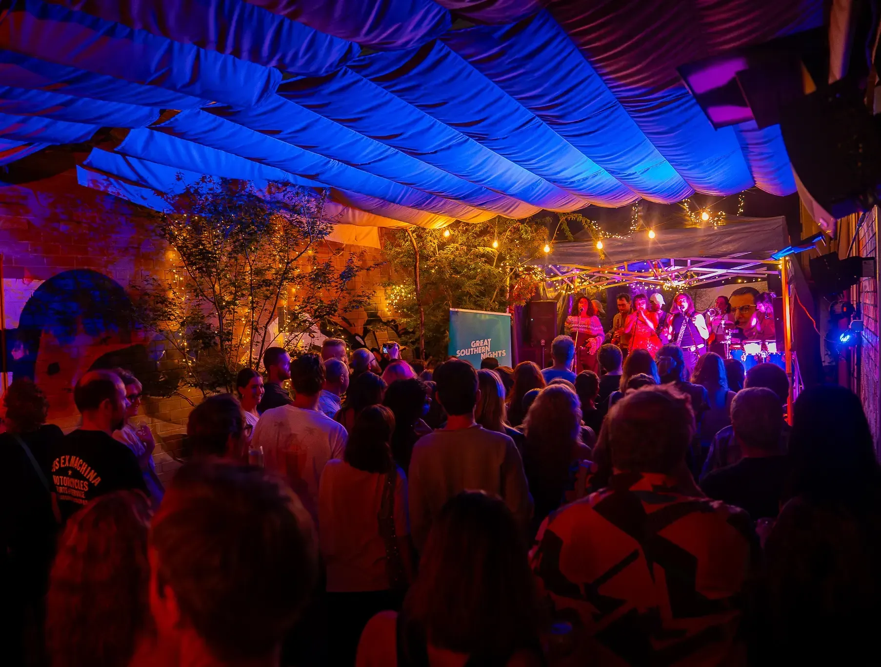 A lively crowd watches a band perform on a colorful, illuminated stage under a canopy with blue fabric and string lights at night.