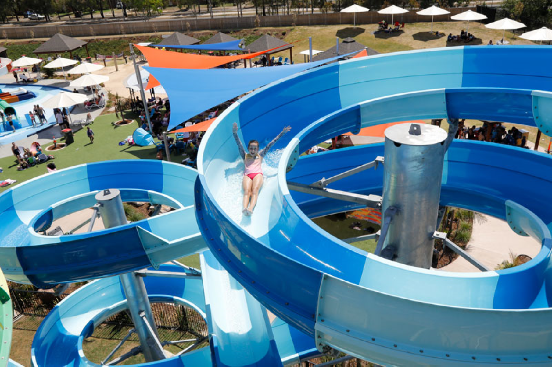 A person in a swimsuit slides down a large, twisting blue water slide at a water park on a sunny day. Umbrellas and people are visible below.