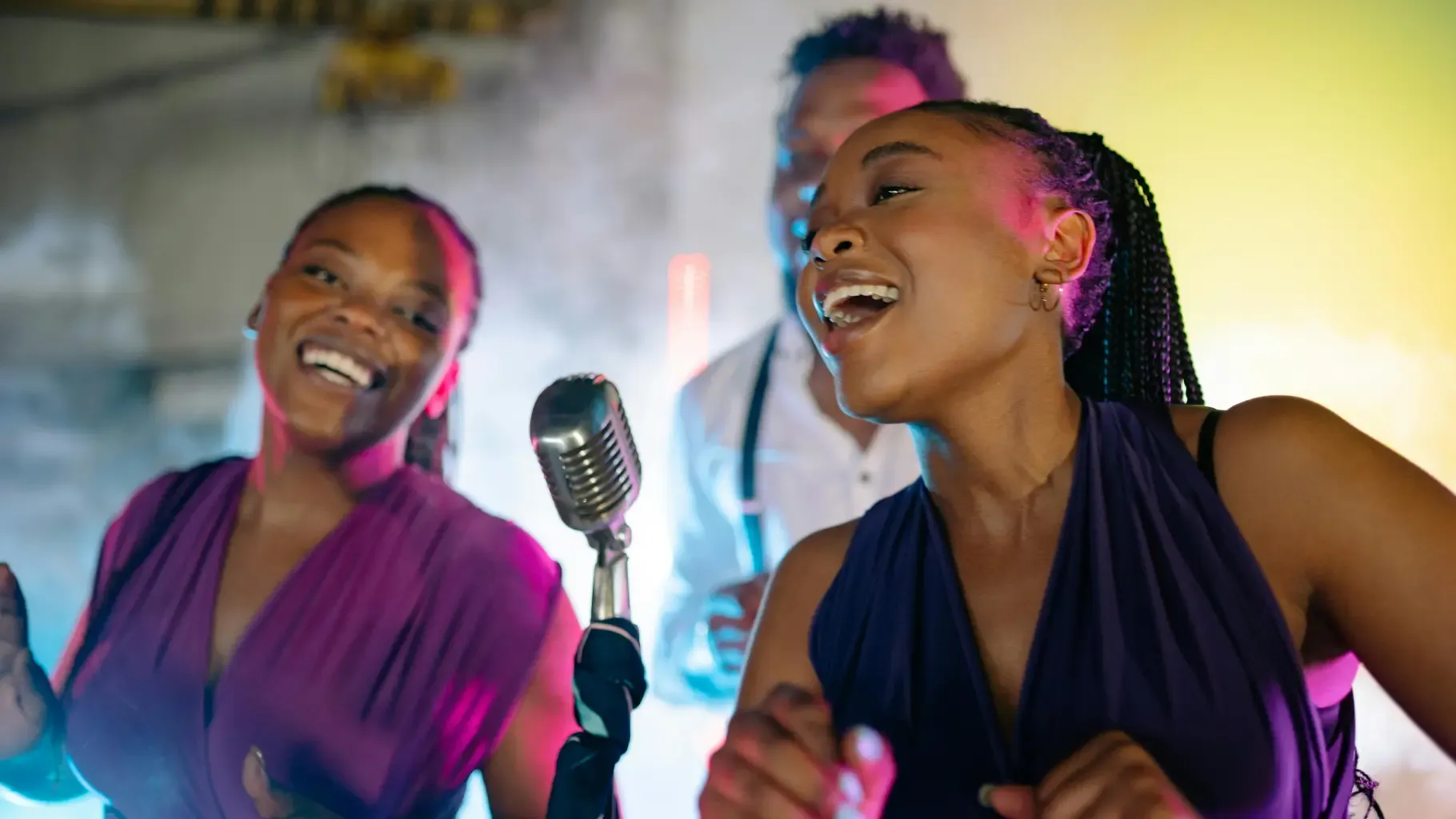 Three people joyfully singing into a vintage microphone on stage, with colorful lights and smoke creating a lively atmosphere.