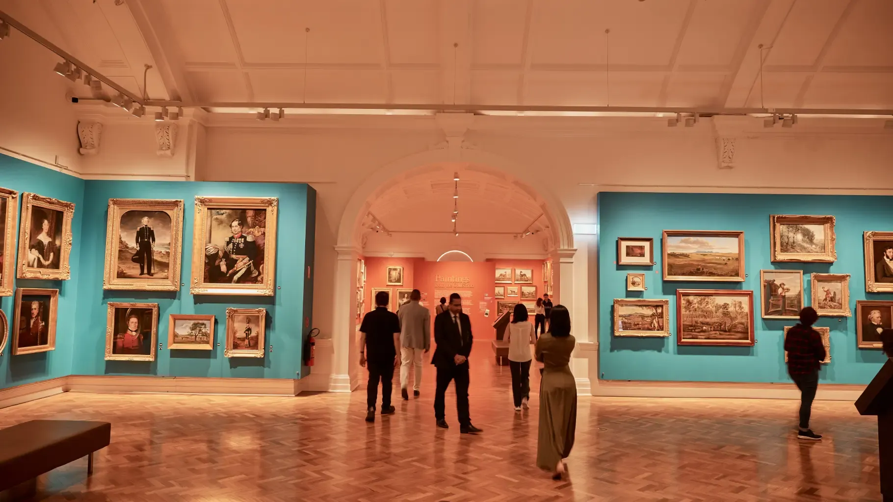 People admire paintings in a spacious art gallery with blue and peach walls, featuring various framed artworks and a wooden floor.