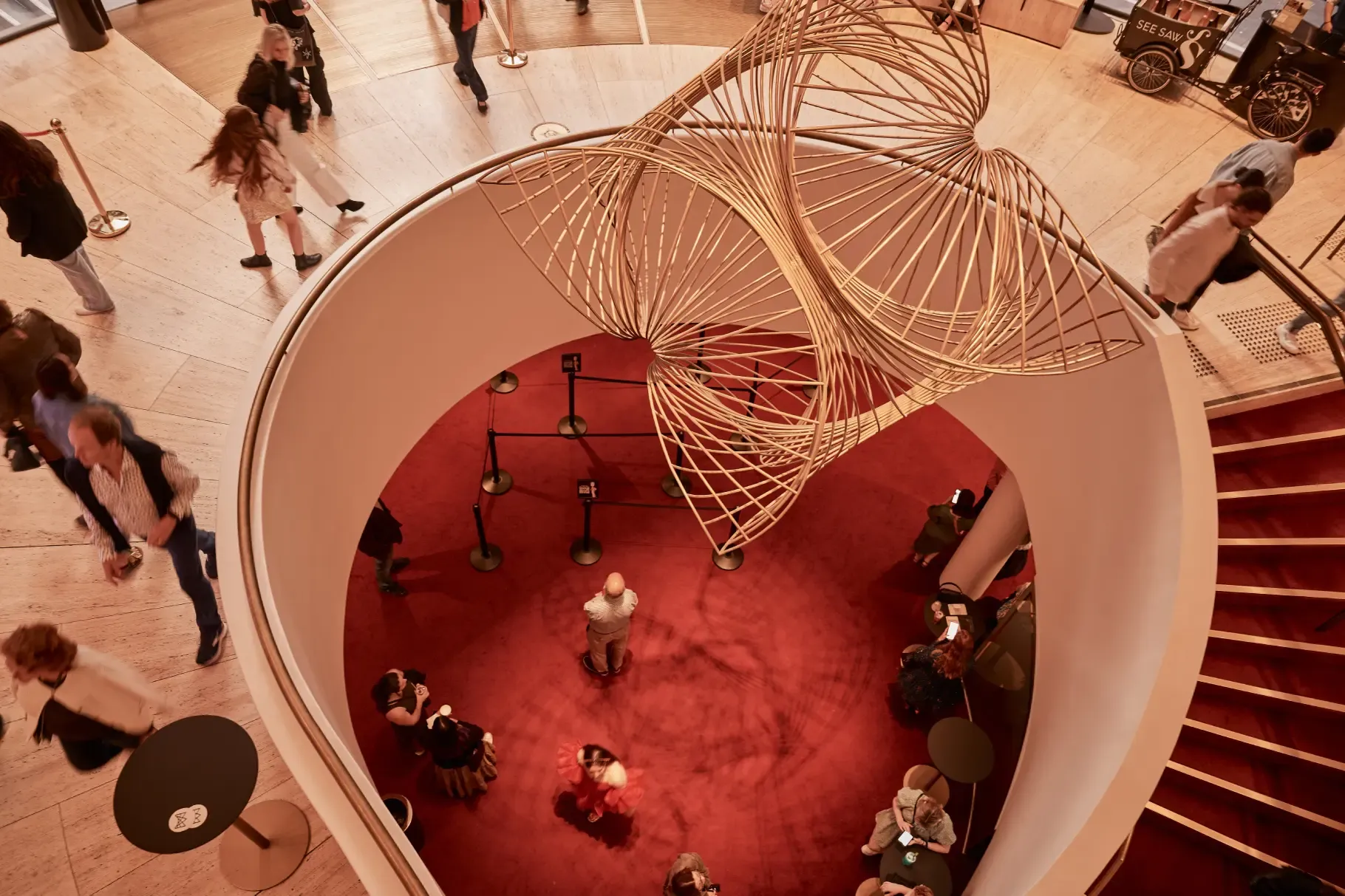Aerial view of a spiral staircase with a large, intricate wire sculpture overhead. People are walking and sitting on red carpeted floors below.
