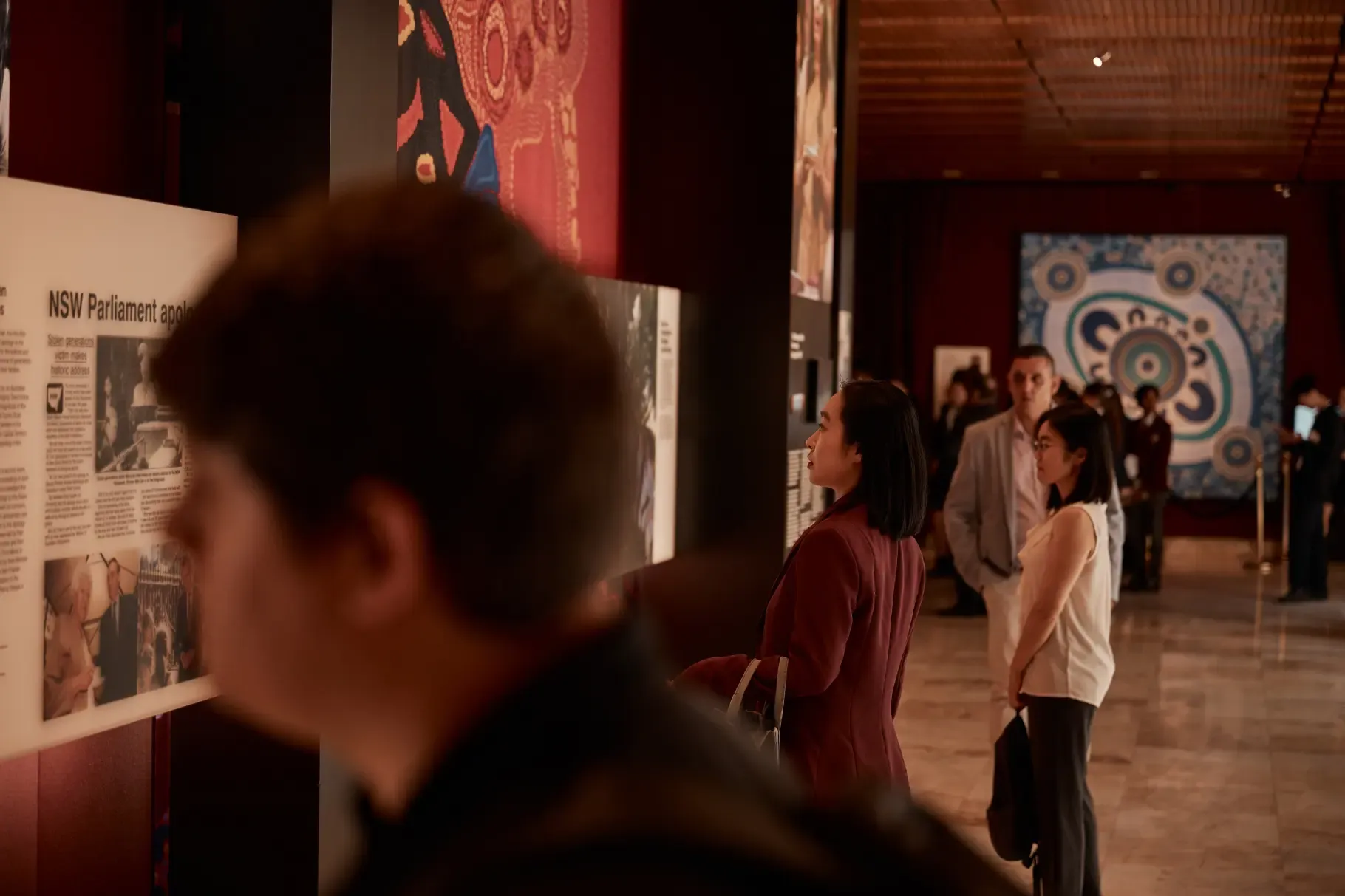 People viewing art exhibits in a dimly lit gallery with colorful abstract paintings on the walls.