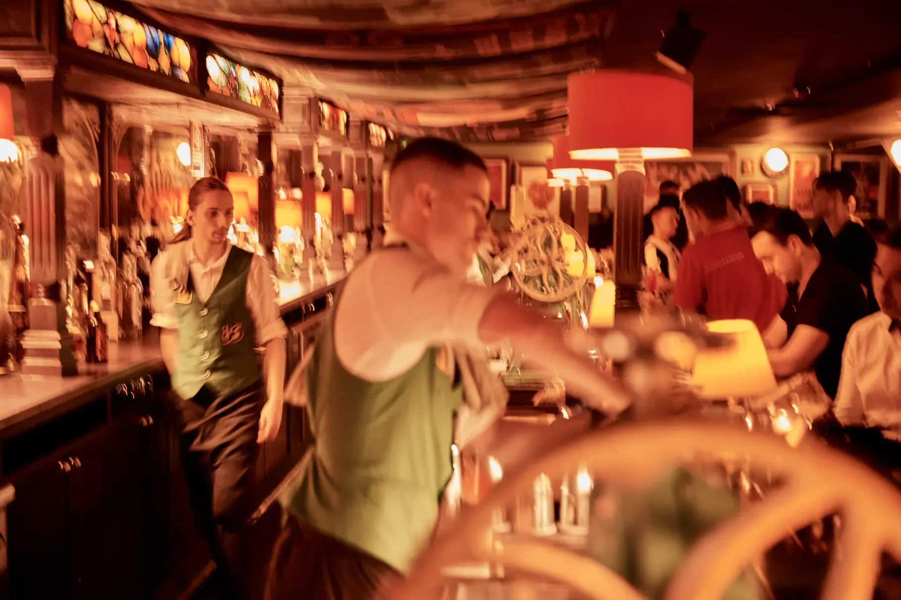 A busy bar scene with bartenders in green vests serving drinks, warm lighting, and patrons seated at the counter.
