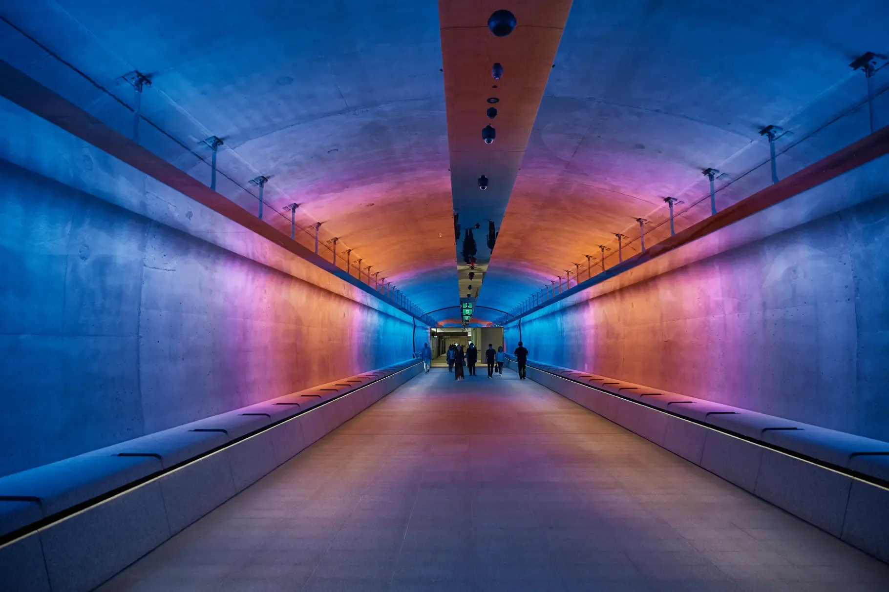A tunnel with vibrant blue, pink, and orange lighting, featuring silhouettes of people walking toward the illuminated exit.