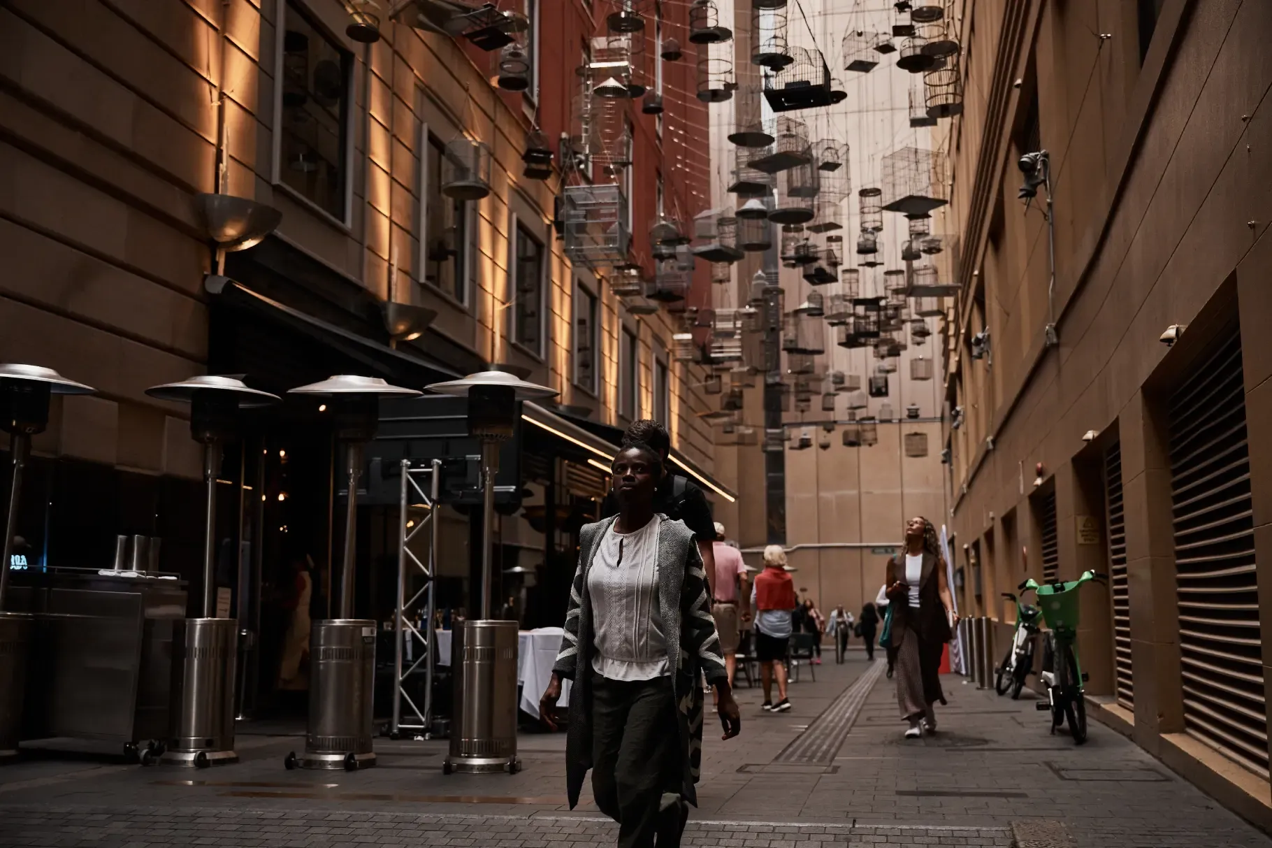 A person walks through a narrow alley with hanging art installations above. The alley is lined with buildings and outdoor heaters.