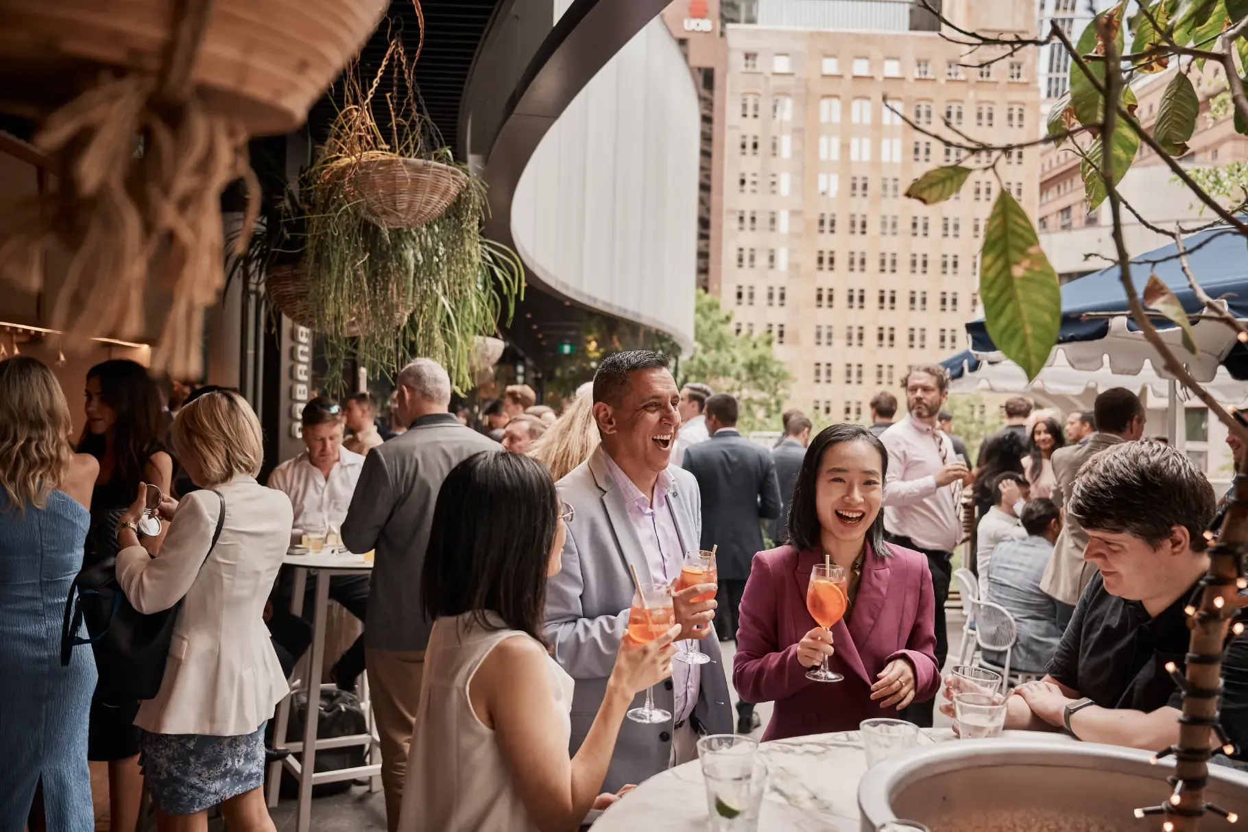People socializing at a lively outdoor gathering, holding drinks and chatting, with city buildings in the background.