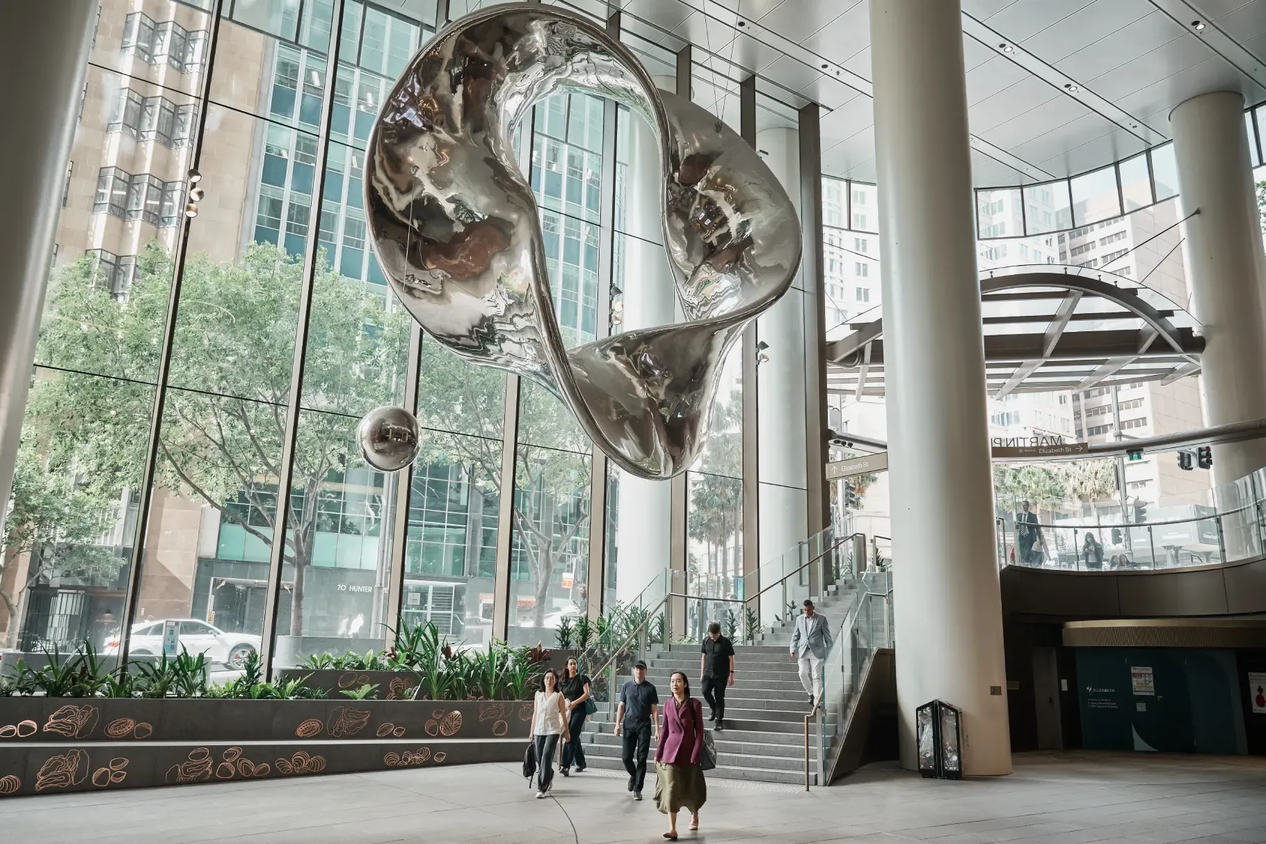 People walk through a spacious, modern indoor area at Martin Place Metro station in Sydney, featuring floor-to-ceiling windows, large white columns, and a suspended mirrored sculpture.