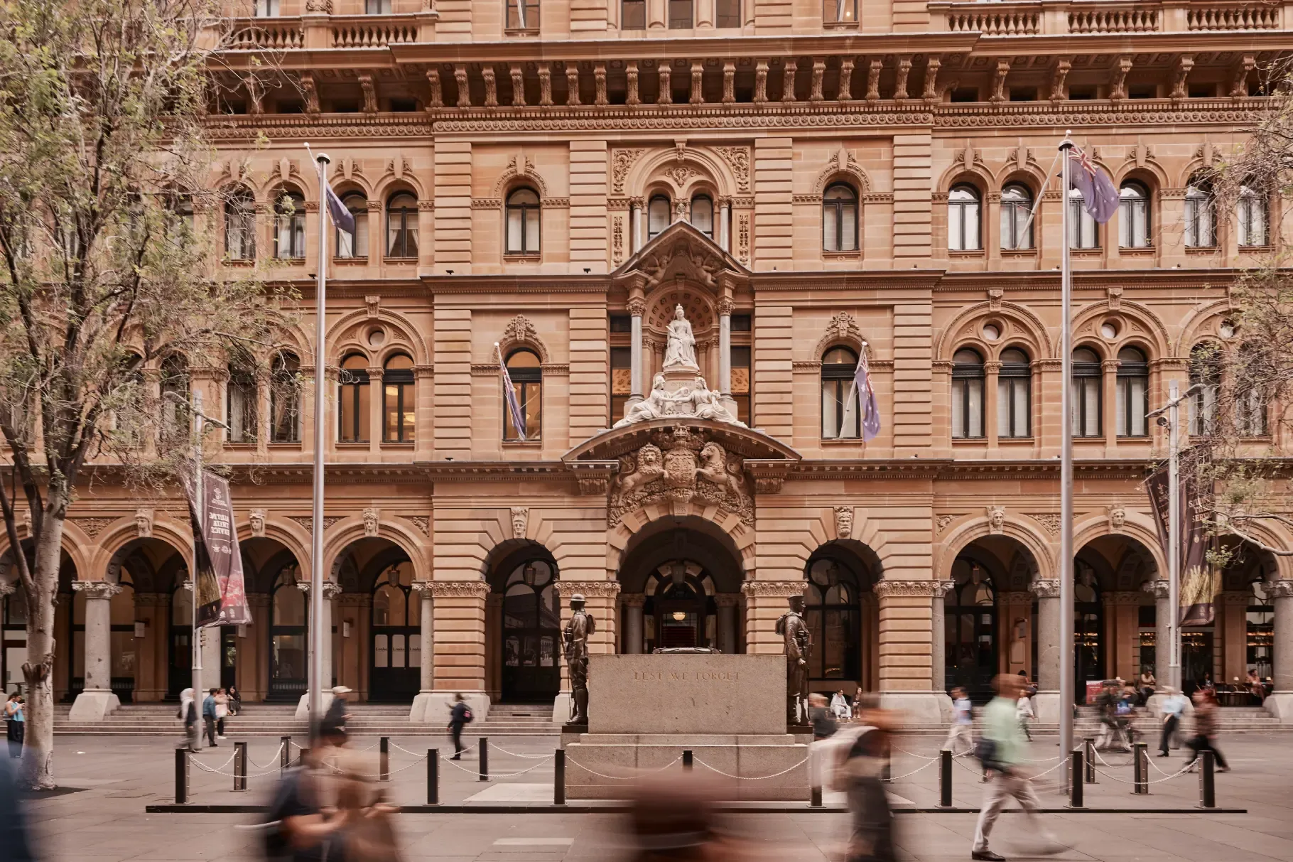 People walk past the historic General Post Office (GPO) building on Martin Place in Sydney, with its grand sandstone facade, arched colonnade, and war memorial featuring two soldier statues