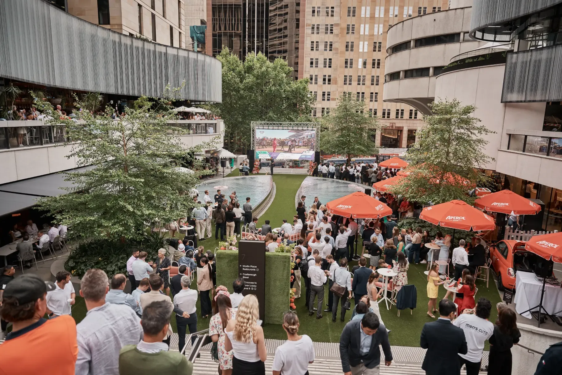 A bustling outdoor event with people gathered around a large screen, surrounded by trees and buildings. Orange umbrellas provide shade.